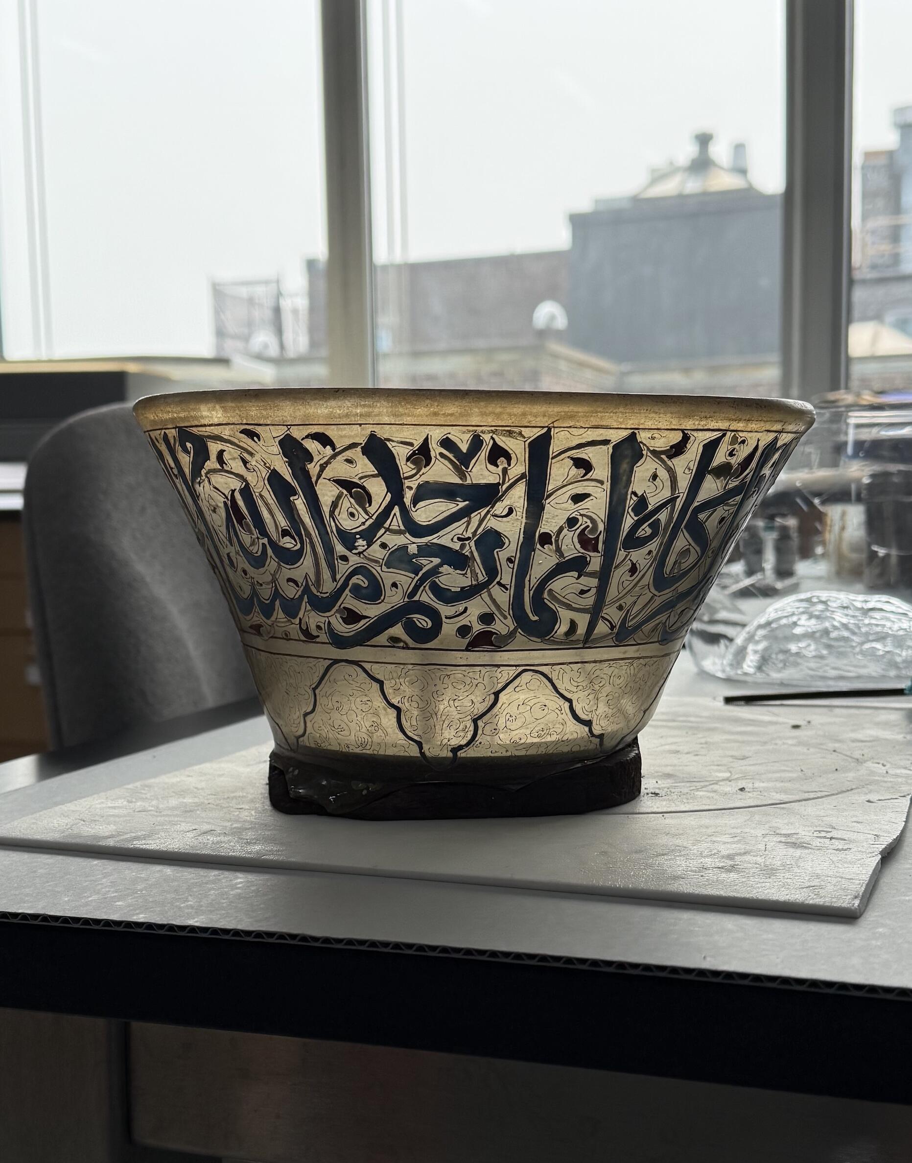 The Fragment of a Mamluk Lamp sits on a table in the conservation lab, supported by balsa painted dark brown in the approximate shape of the lamp’s break edge and two acrylic disks on the interior. Light from the windows shines through the translucent glass.