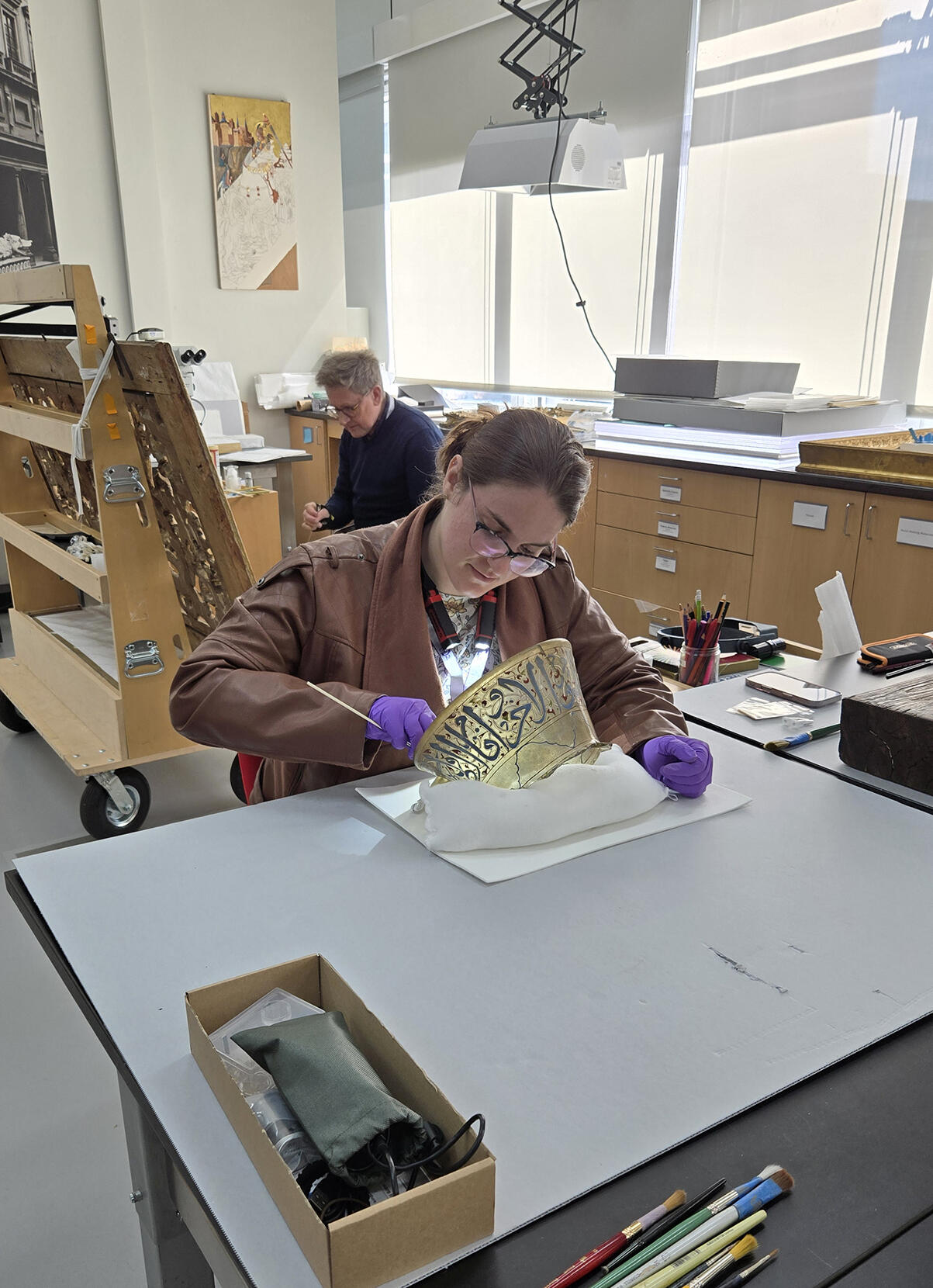 A white woman with brown hair pulled back, glasses, and a brown jacket sits at a table in the Conservation Lab, cleaning the interior of the oil  lamp fragment.