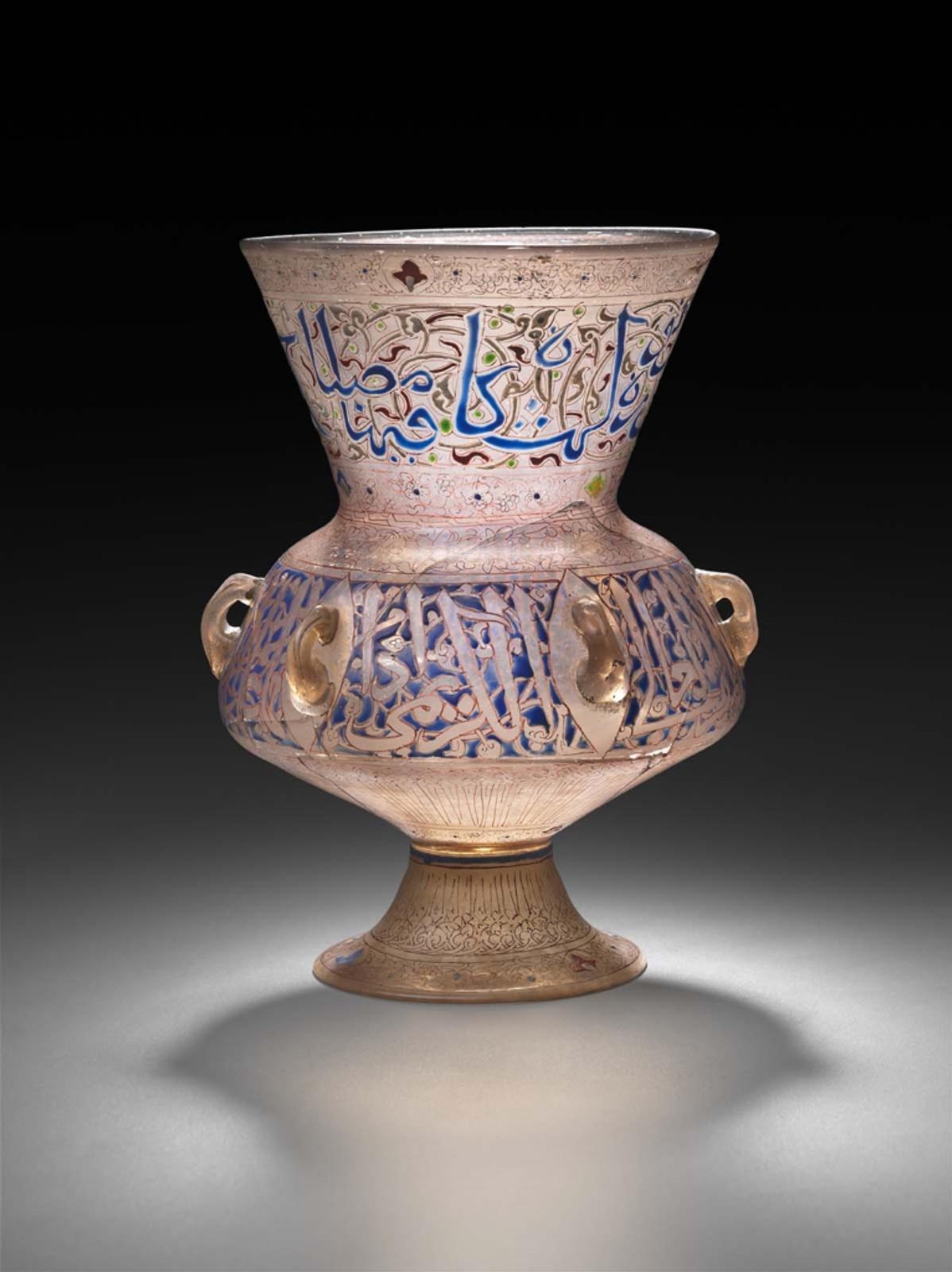 A complete enamelled glass  mosque lamp with a form that tapers into a neck then flares out to create a rounded belly. Blue Thuluth script encircles the neck and clear Thuluth script outlined in blue enamel encircles the belly.