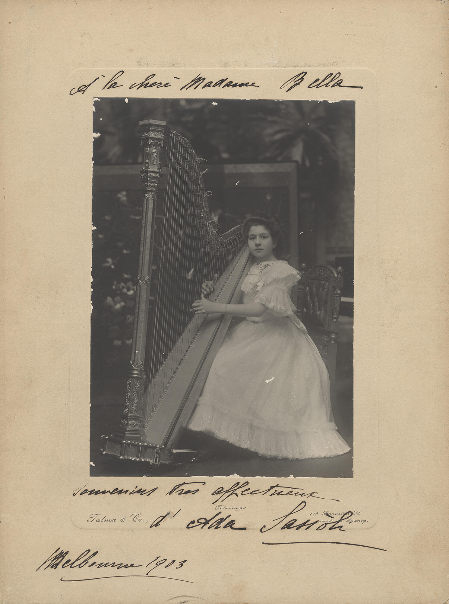 A black and white photograph of a white woman seated on a wooden chair, playing a harp.