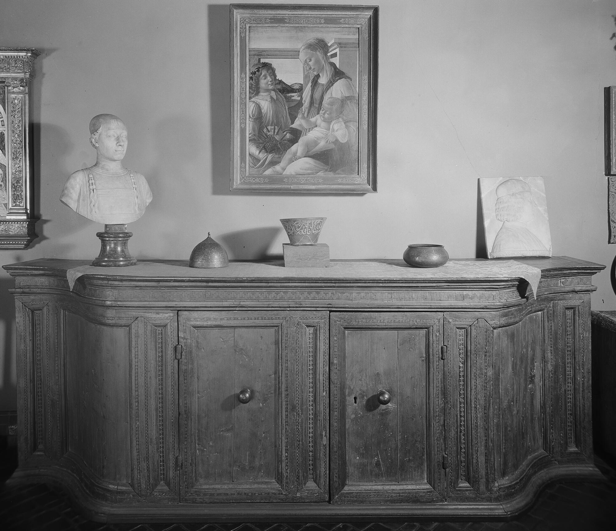 A black and white photograph of a flared neck and rim of a glass oil lamp on top of a wooden credenza flanked by an Islamic metal bowl and helmet. The oil lamp fragment is translucent there is no white support at its base.