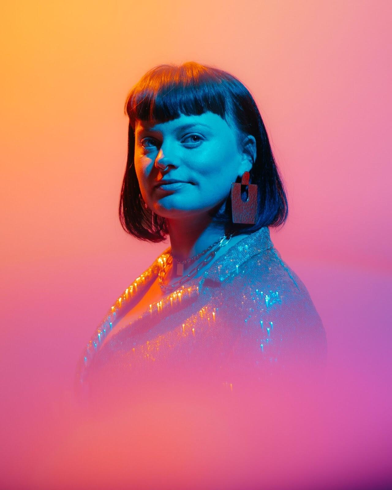 Headshot of artist with short dark hair and bands in a orange pink gradient background 