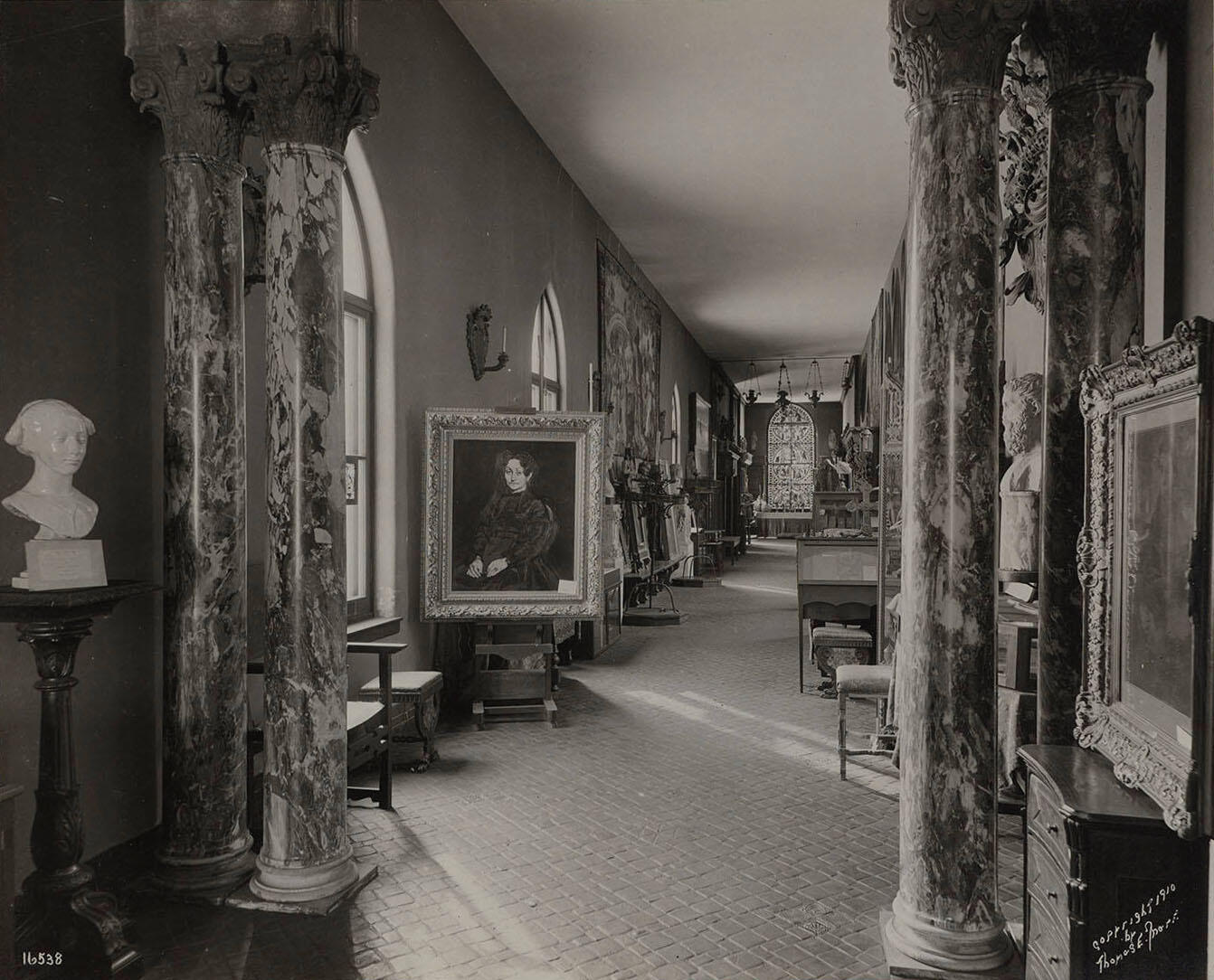 A black and white photograph of a long, narrow gallery filled with artwork, furniture, and a large stained glass window.
