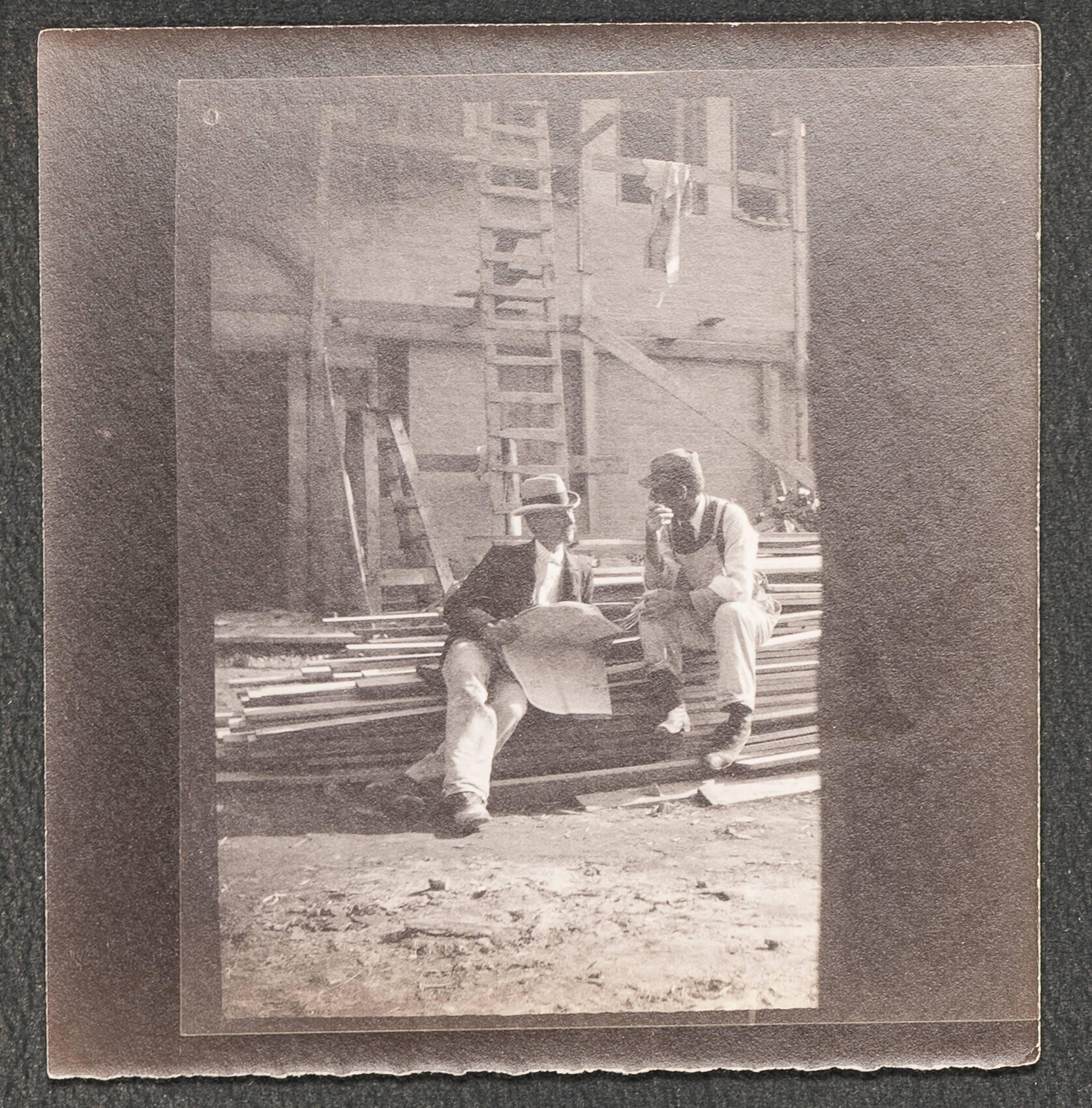 A black and white photograph of two white men sitting in a pile of lumber on a construction site.
