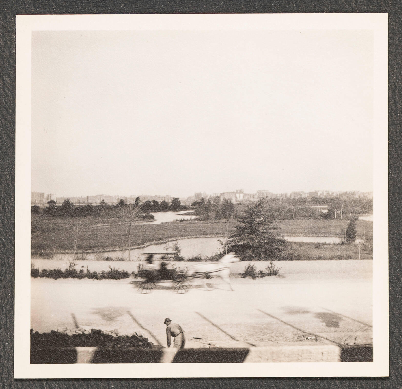 A black and white photograph of a horse-drawn carriage passing by in front of the Muddy River with the Boston skyline in the distance and a construction laborer in the foreground.