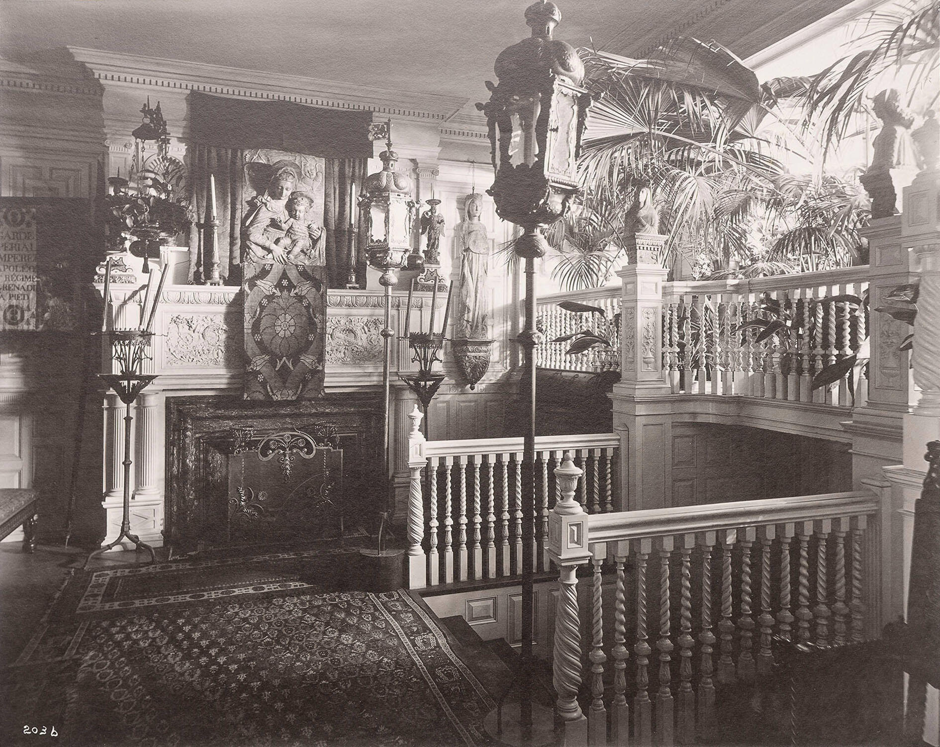 A black and white photograph with a room filled with carved woodwork and paneling, artwork, textiles, and plants.