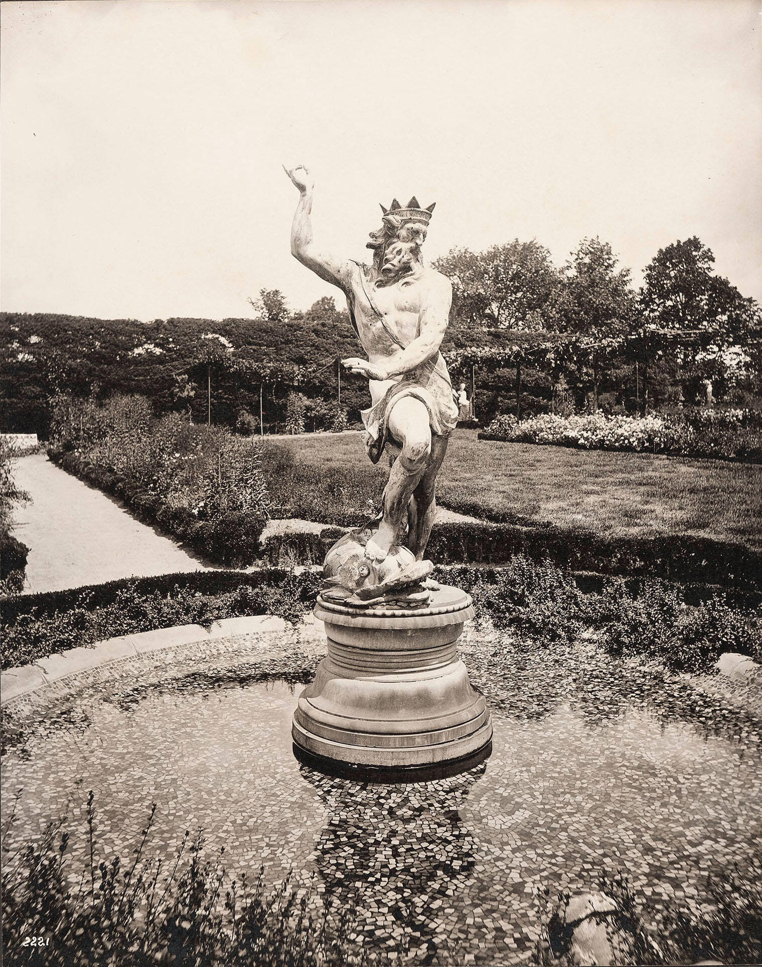 A black and white photograph of a circular fountain with the figure of Neptune in a garden. 