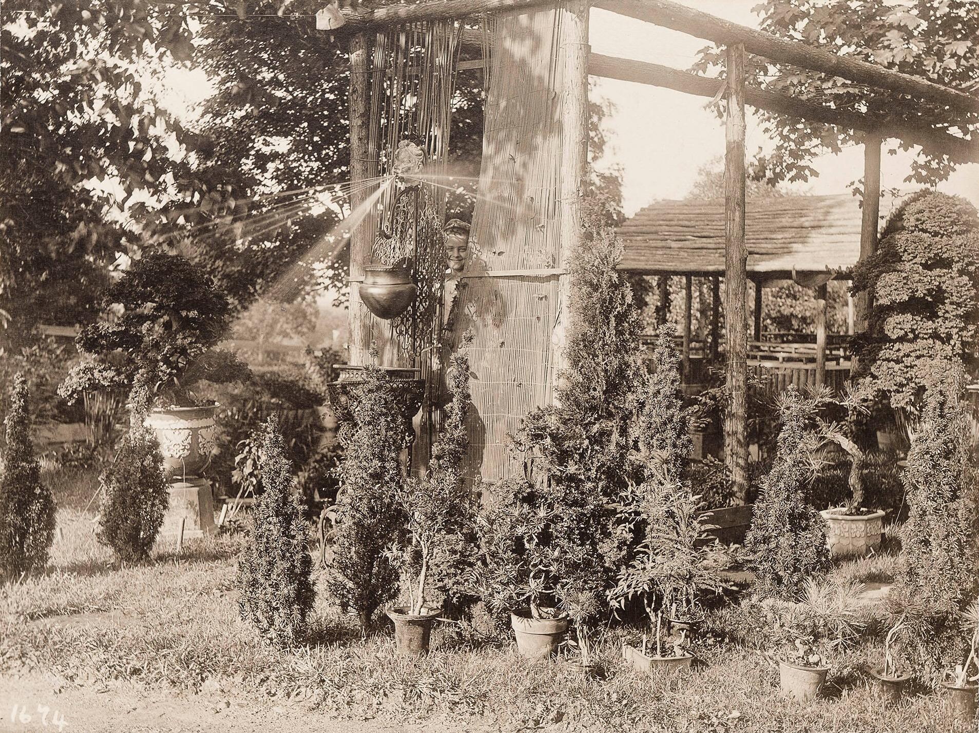A black and white photograph of a garden with a wooden frame, potted small trees, and fountain spraying water.