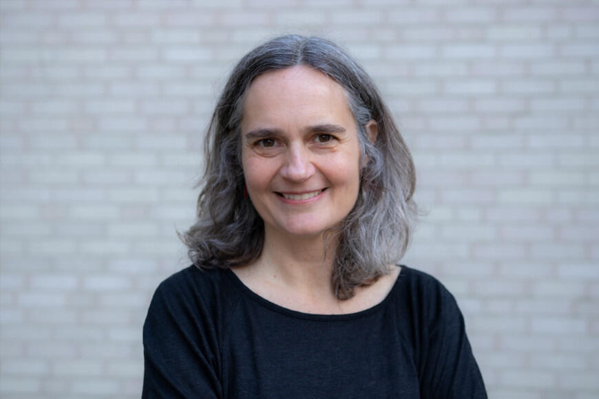 woman with short gray hair wearing a black top standing in front of a white brick walla