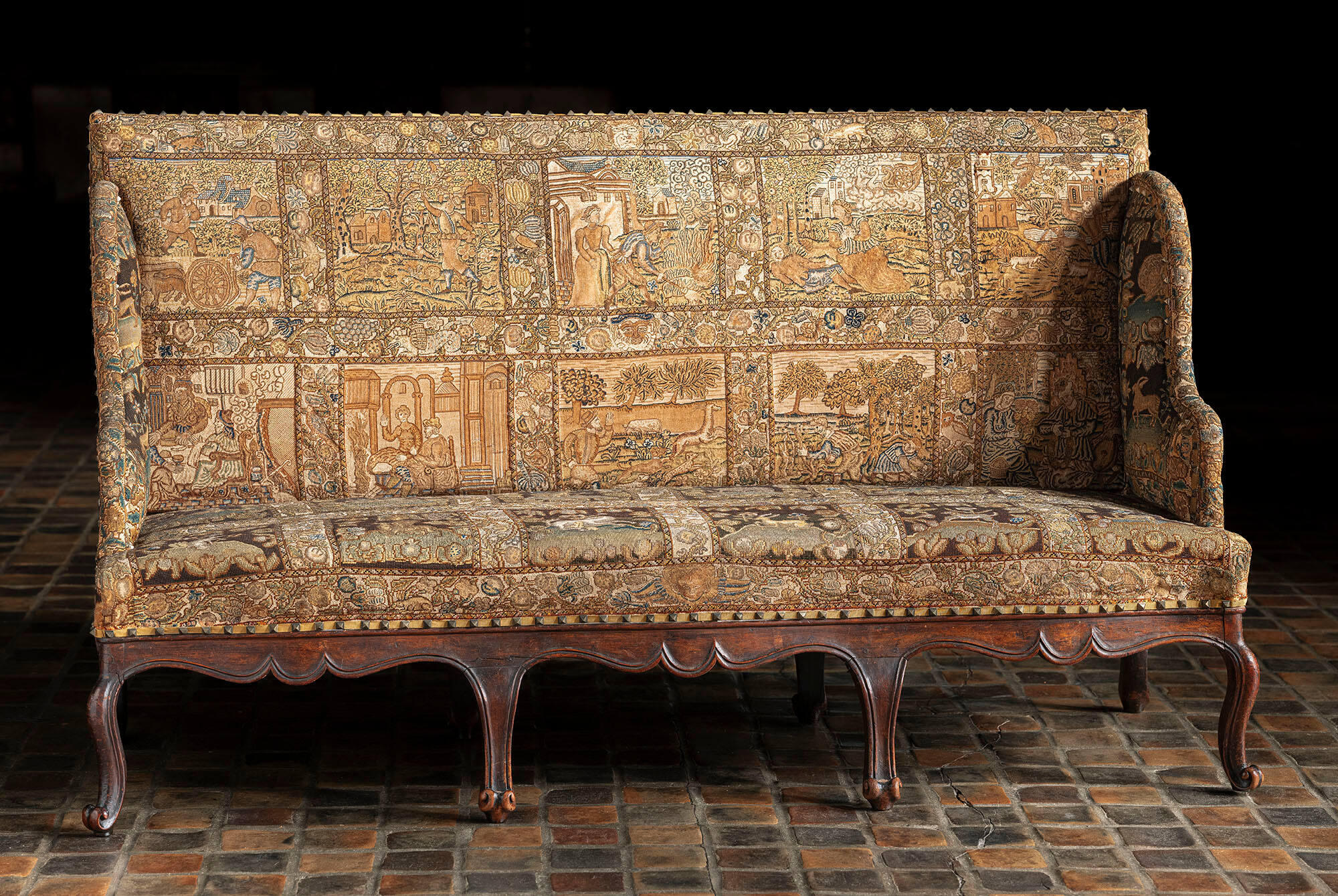 A sofa with a high back and detailed embroidery showing squares of figures in different landscape settings.