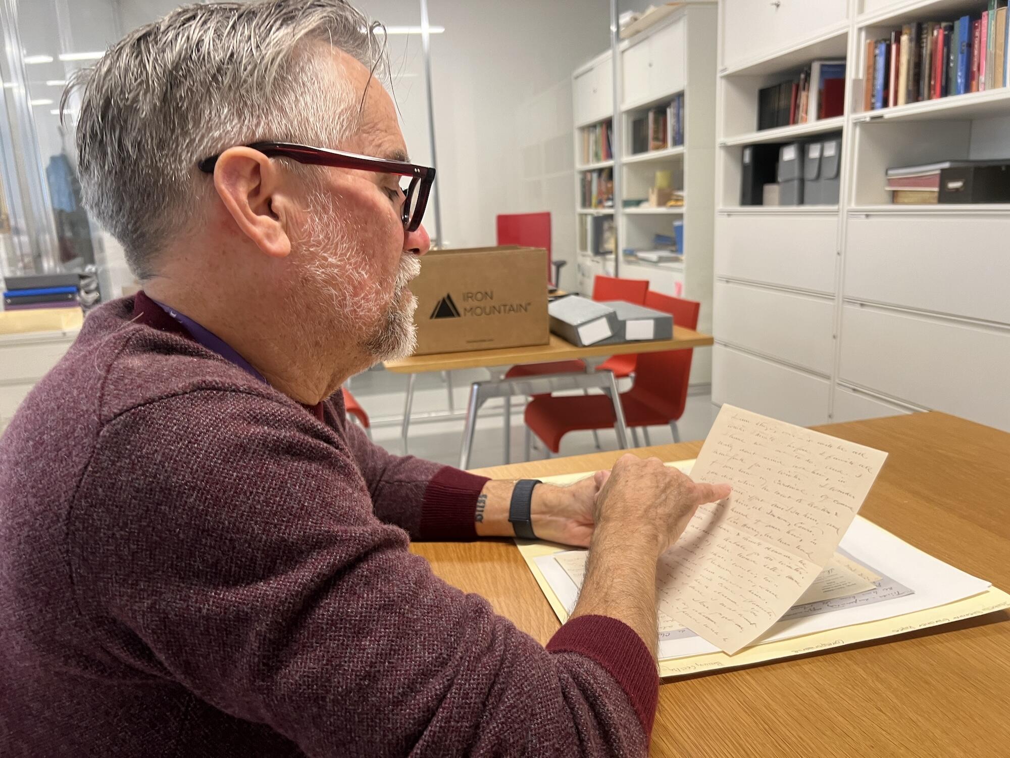 A white man with classes and a red sweater looks at an archival paper covered in handwritten script in a brightly lit room.