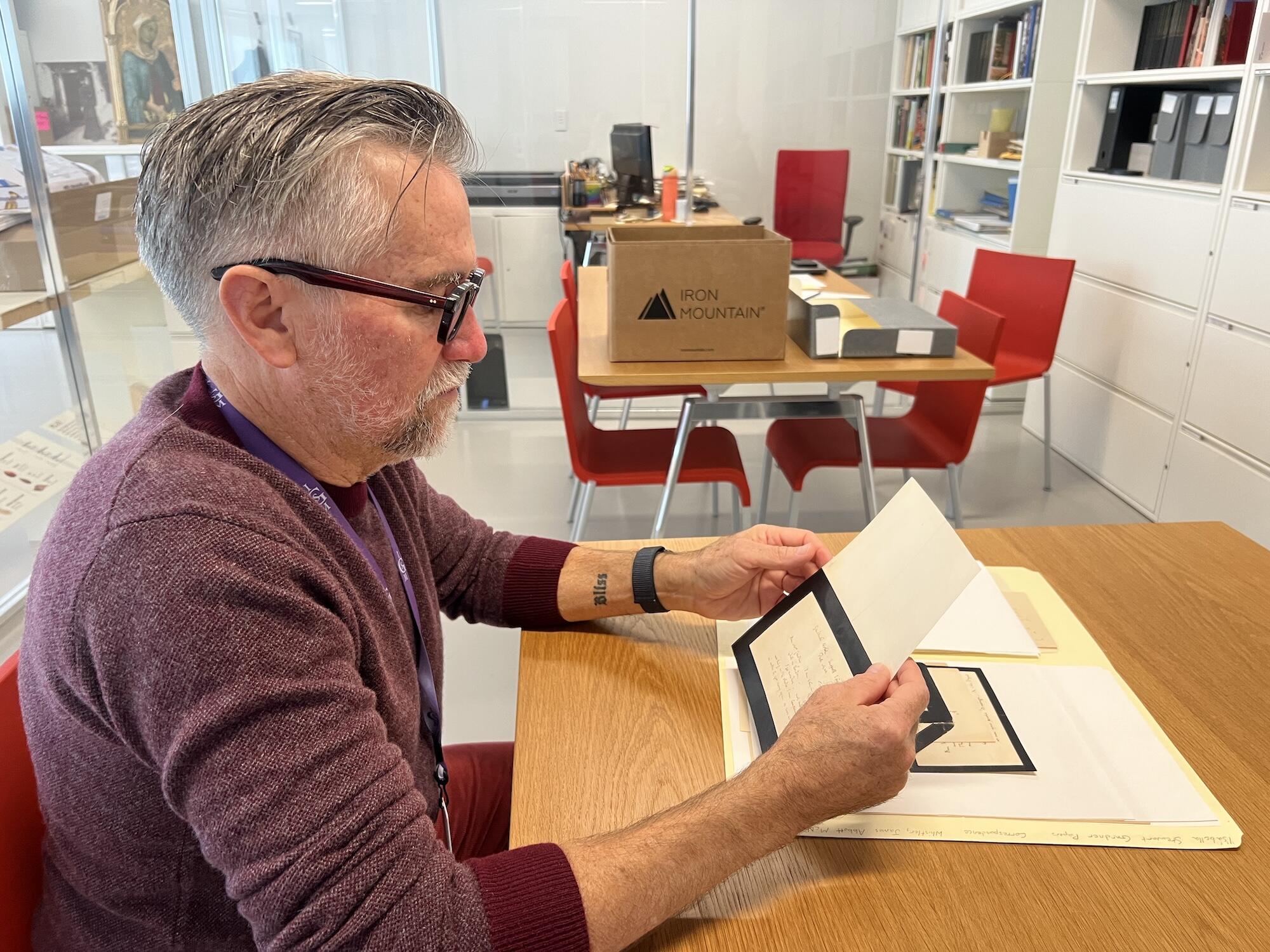 A white man with classes and a red sweater looks at archival papers in a brightly lit room.