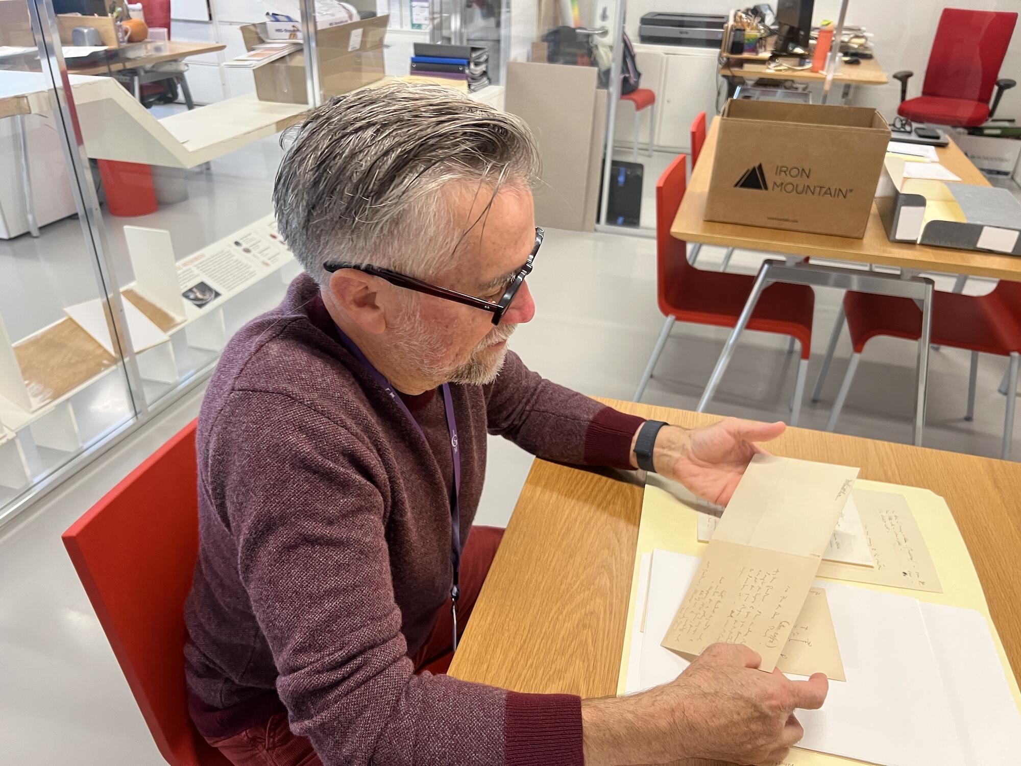 A white man with classes and a red sweater looks at archival papers in a brightly lit room.
