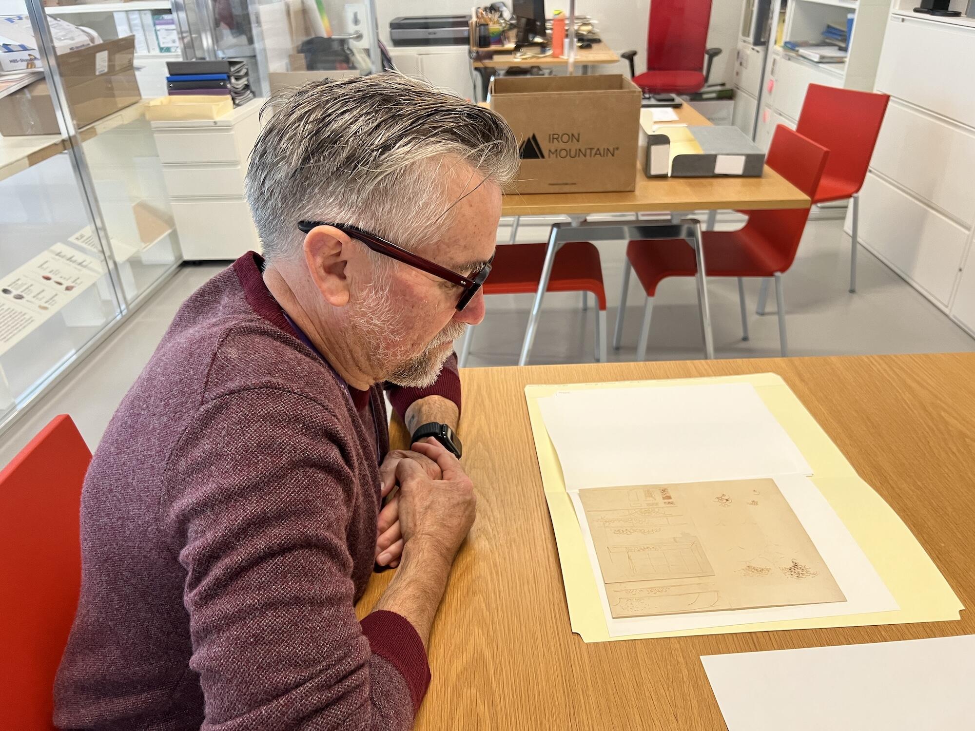 A white man with classes and a red sweater looks at archival papers in a brightly lit room. 
