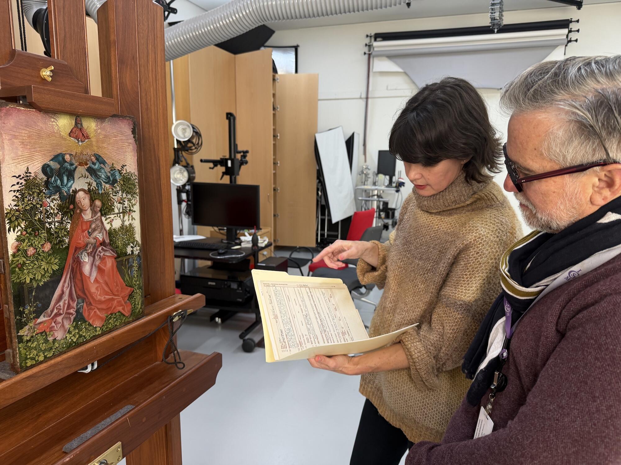 A white man with glasses and a scarf stands next to a white woman with dark hair and a sweater before a painting of the Virgin and Child. The two people look at a manilla folder open to printed notes.