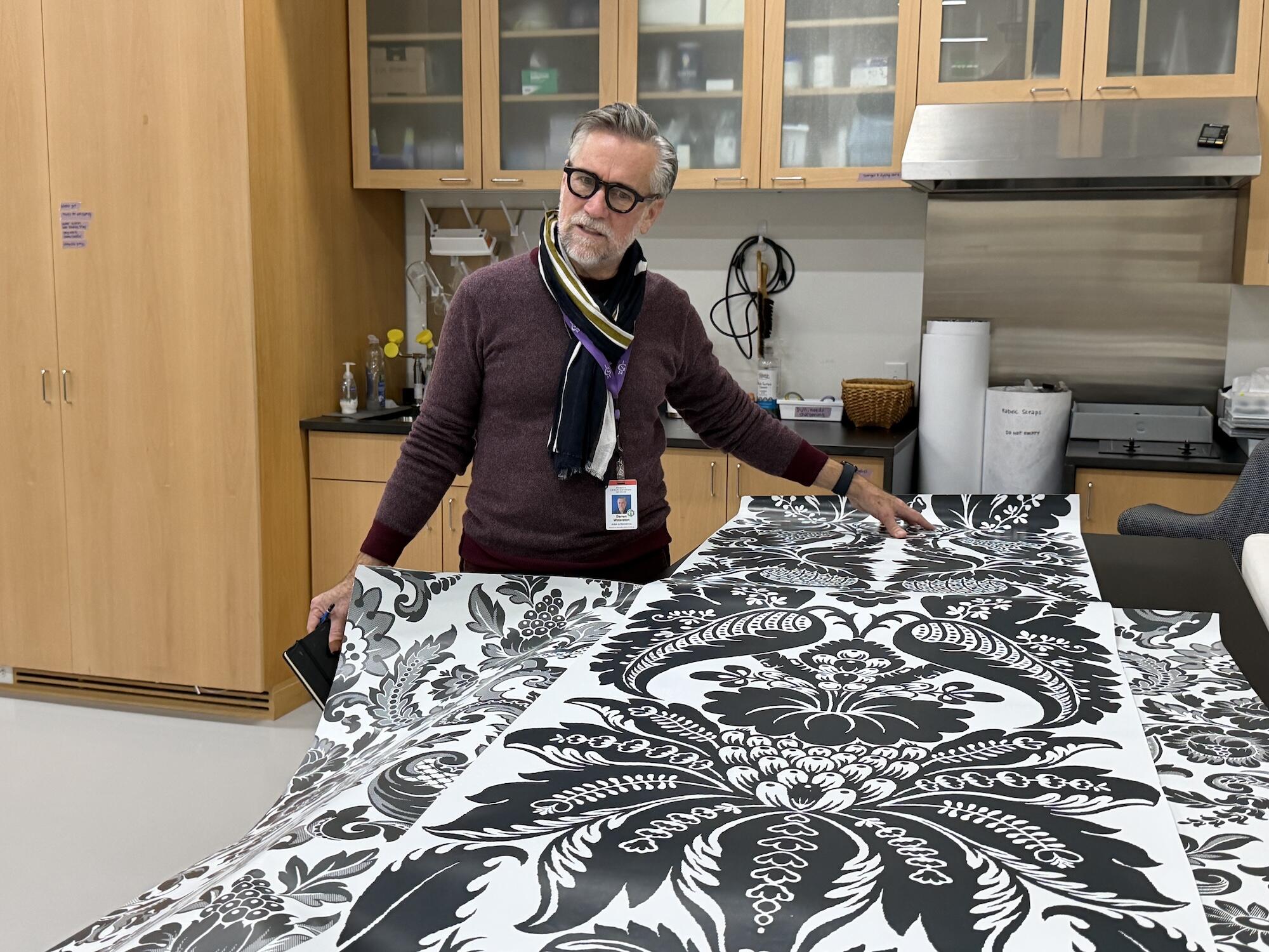 A white man with glasses and a scarf looks at a bold black-and-white print design on long strips of paper. 