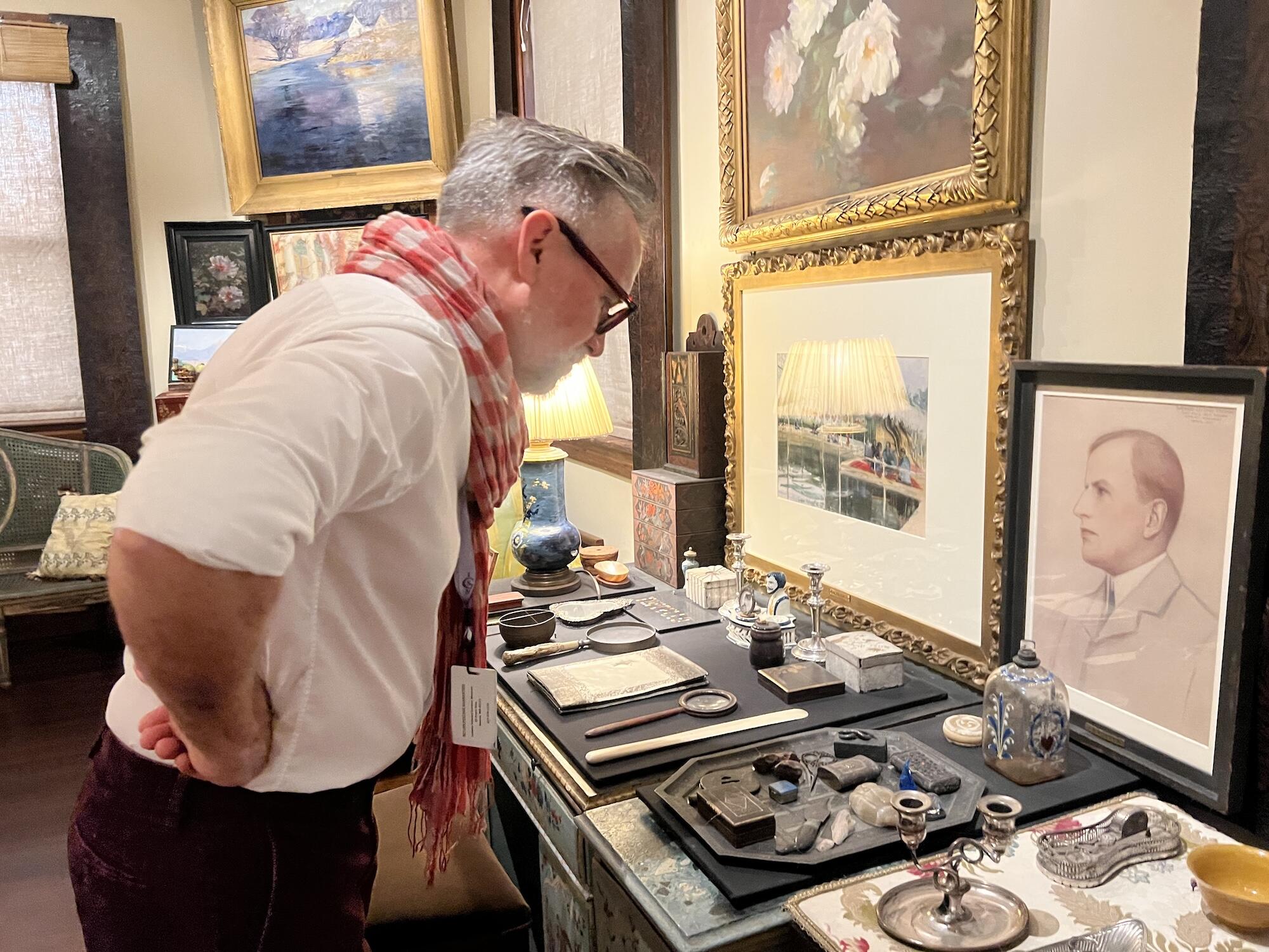 A white man with glasses and a checkered orange scarf leans over a display of small objects displayed on a desk in a room with many framed pictures on the walls. 
