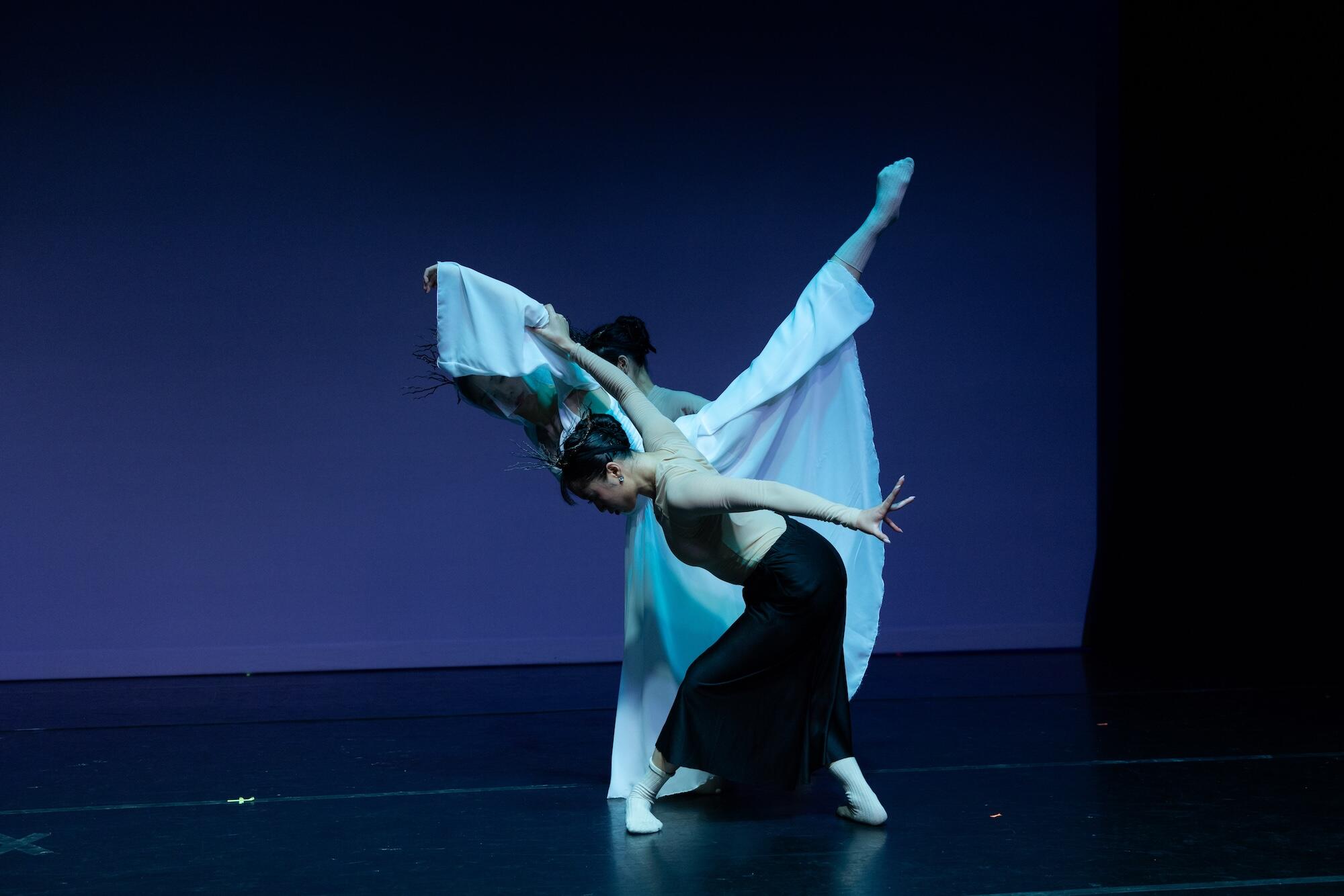 Two dancers on a dim stage. The dancer in front wears a black dress, the dancer behind wears a white robe. The dancers are posed to create an X shape with their bodies.