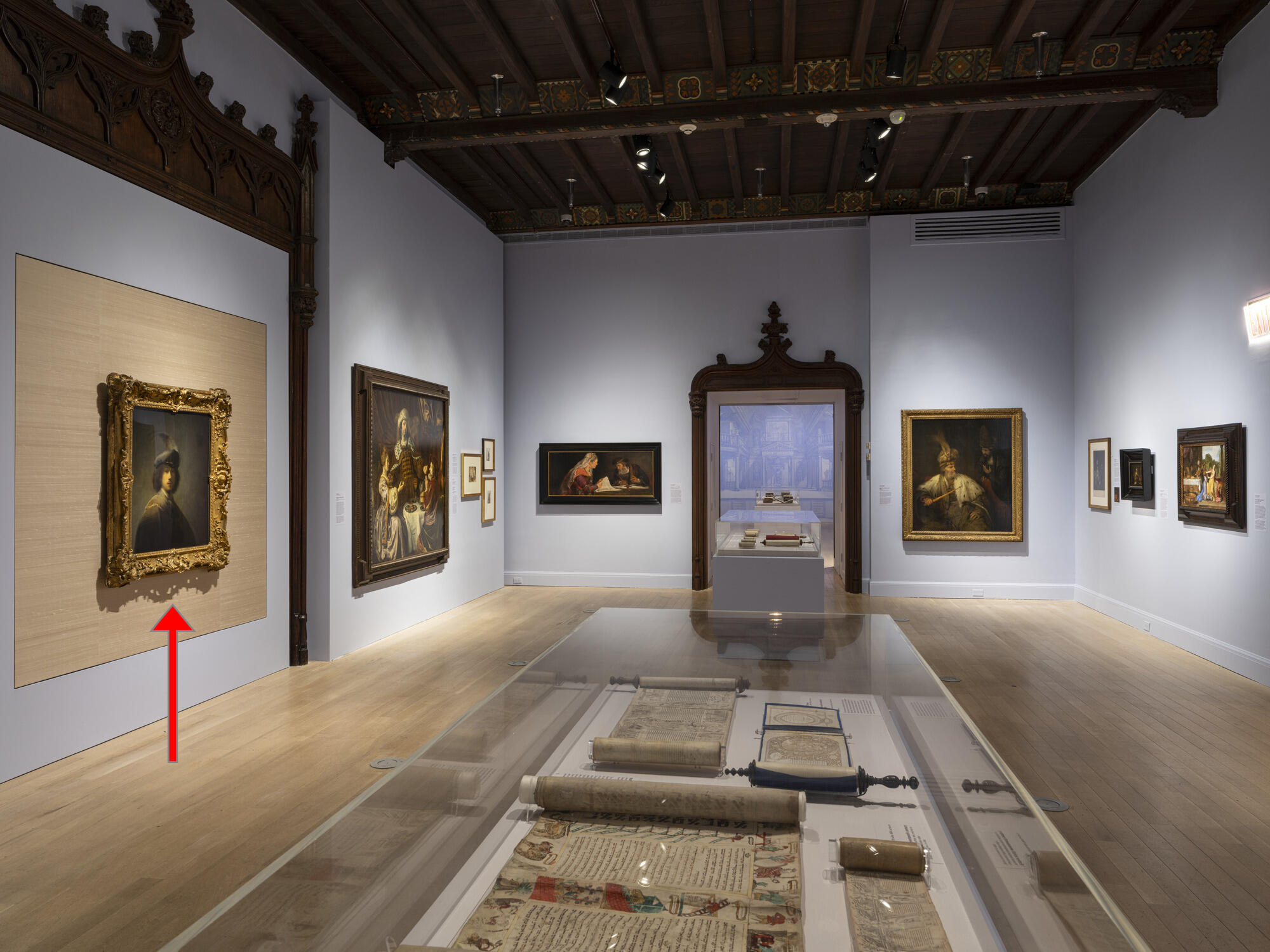 An exhibition gallery with paintings and a display case with scrolls. A red arrow points to a portrait of a young white man