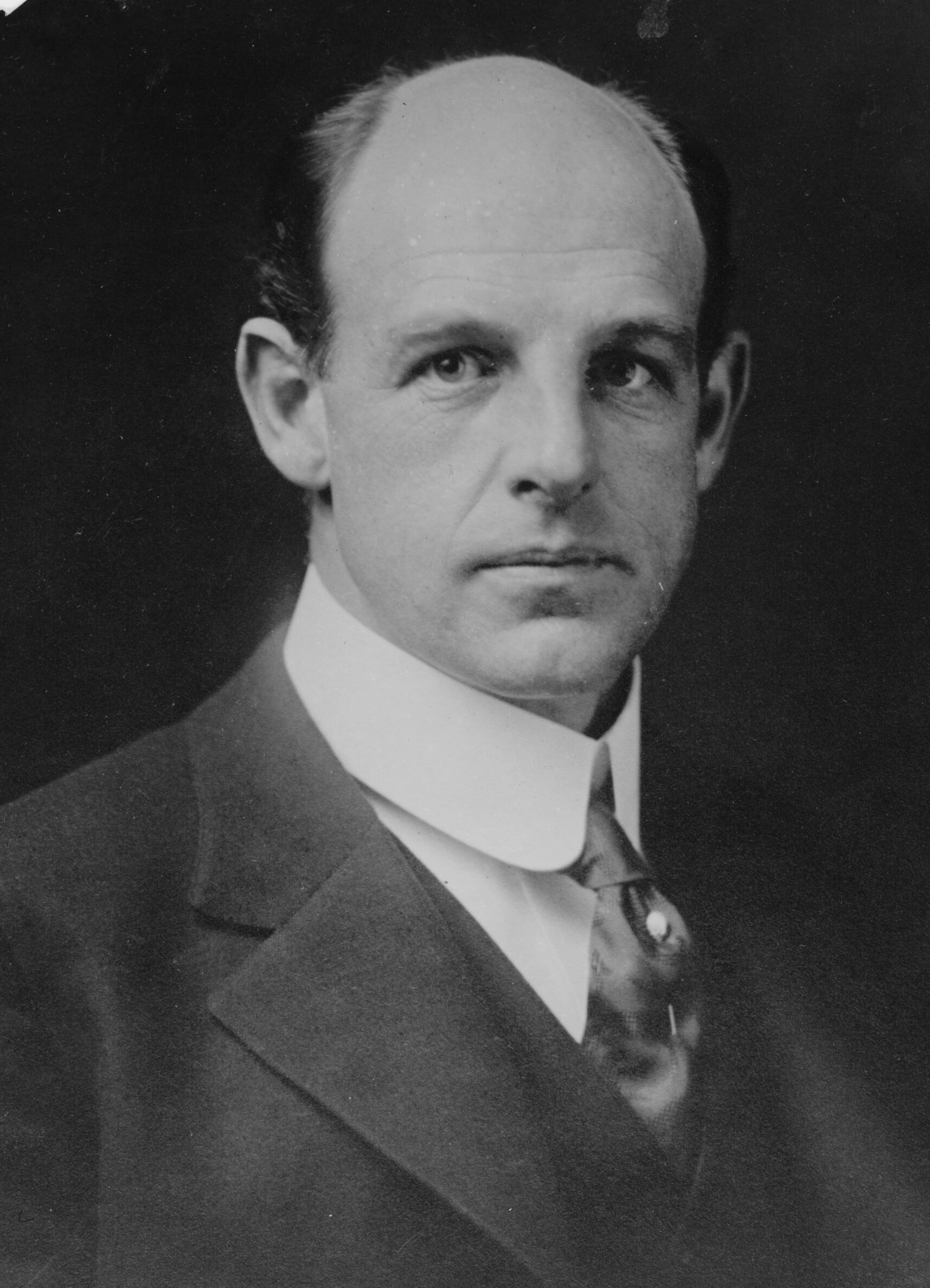 A black and white photograph of a bald white man in an early 20th century suit. https://en.wikipedia.org/wiki/Percy_Haughton