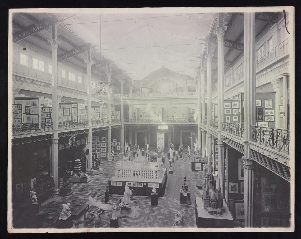 A black and white photograph of a large room with a mezzanine filled with framed artworks, sculptures, and architectural models