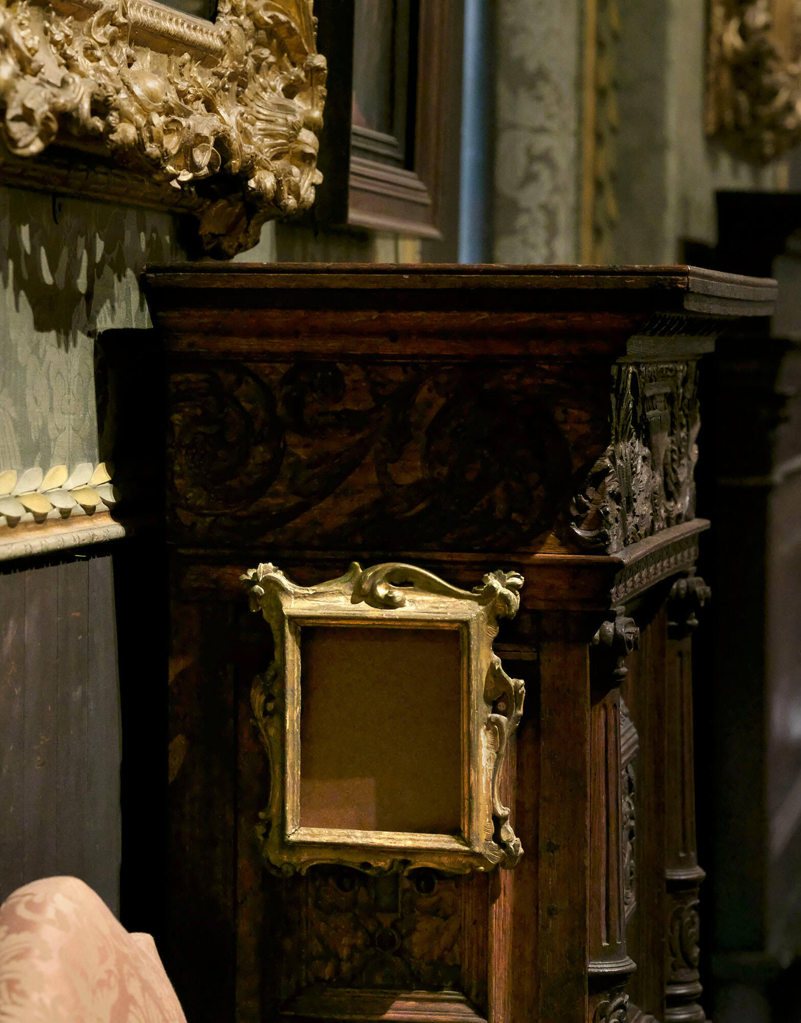 A gilded empty frame hangs on the side of a carved wooden cupboard.