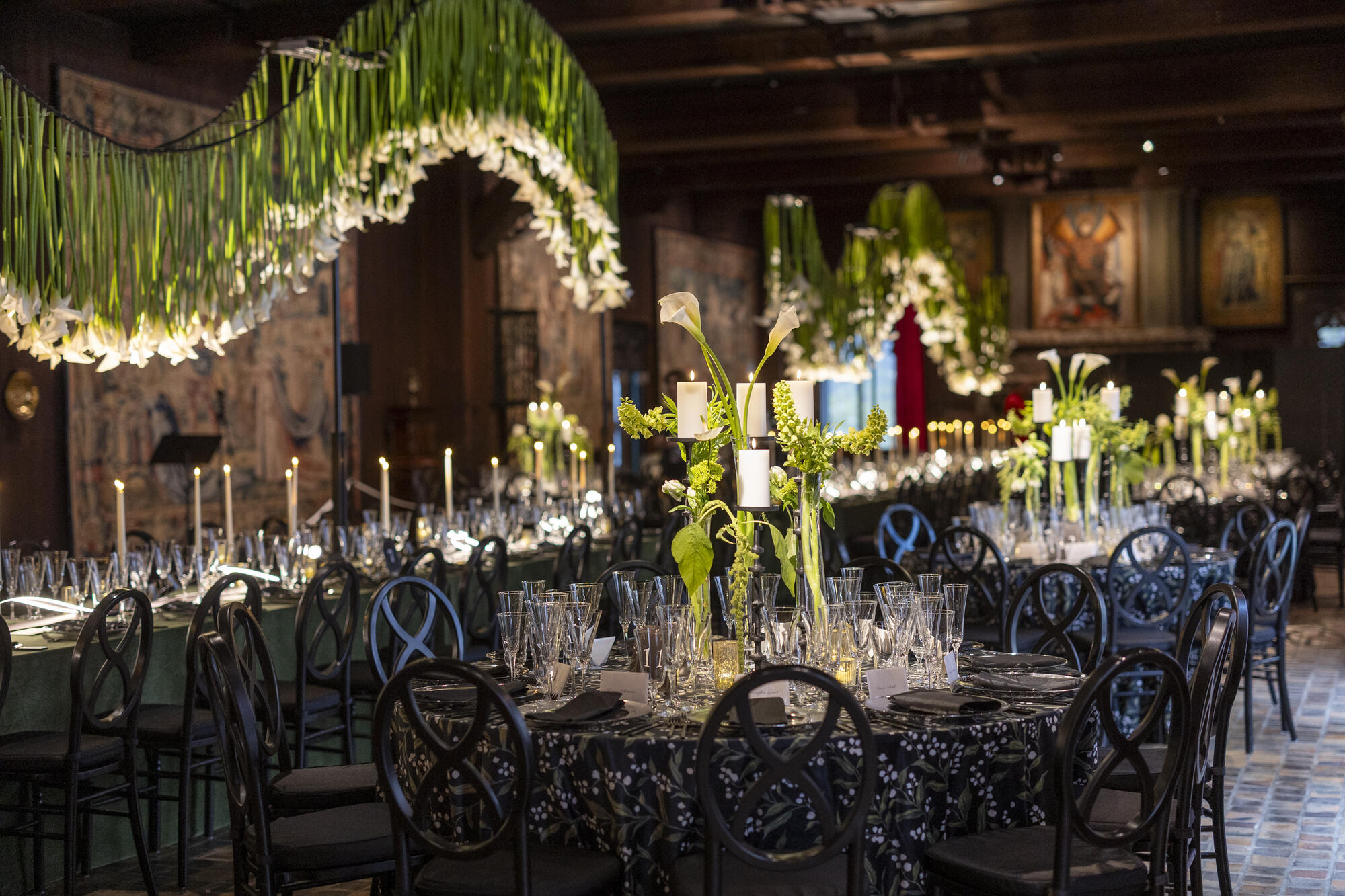 A long table set with glassware and candles with lilies hanging upside down from the ceiling above. 