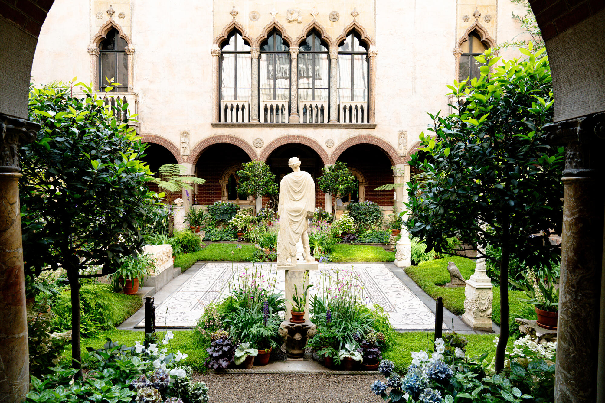 A sunlit courtyard with stone walls, a mosaic floor, and marble statues filled with blooming hydrangeas.