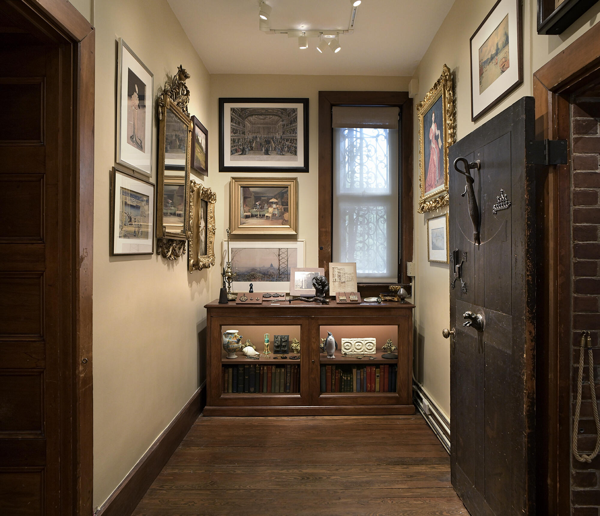 A narrow room with a bookcase filled with books and objects and paintings on the wall.