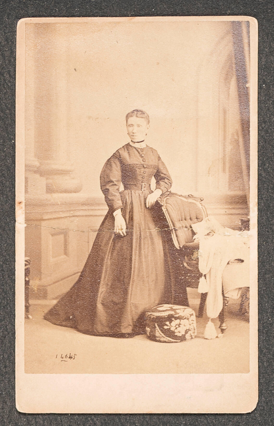 Full-length photograph of a light-skinned woman wearing a dark, long-sleeved dress. She is photographed in a studio, with her arm resting on a chair and architectural imagery in the background.