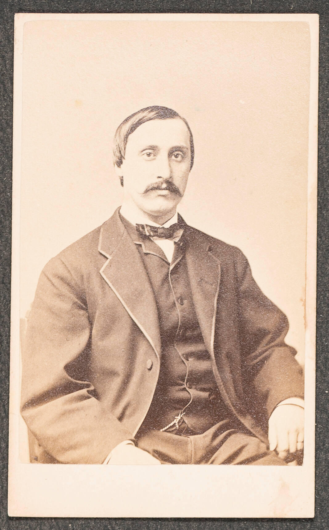 A brown-toned photographic portrait of a seated man from the waist up. He has light skin, dark hair slicked down and a mustache, and is wearing a dark suit with a light shirt and dark bow tie. 