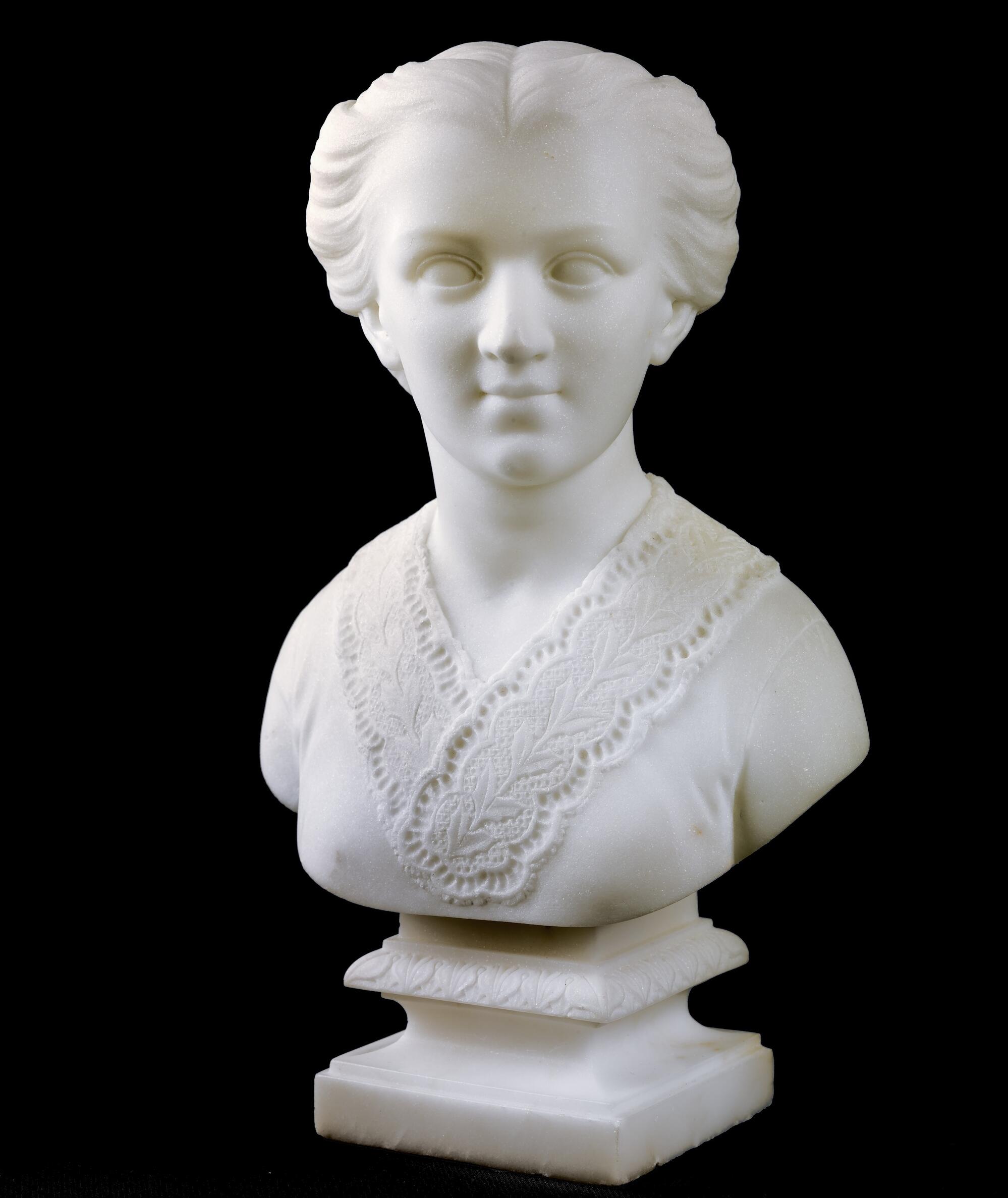 Sculpture bust in marble of a girl with a slight smile, her hair pulled back, and wearing a v-neck garment with lacy trim.