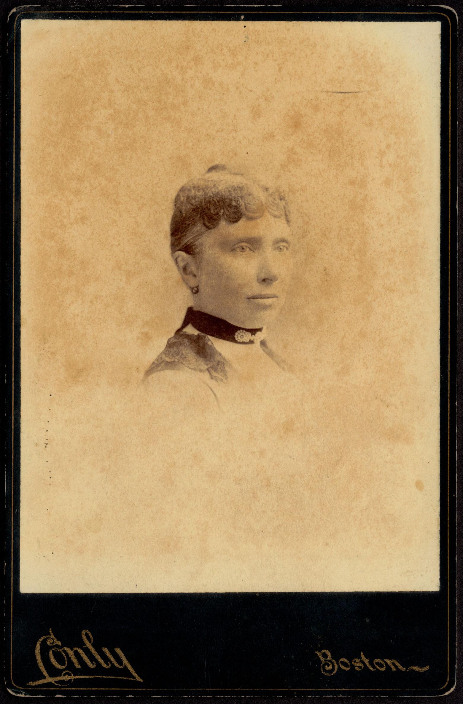 Photograph of a woman from the shoulders up, with light skin and medium-dark hair in a tightly curled and flat style. She has a neutral expression, is angled to the viewer’s right, and wears a dress with a dark band collar and lacy shoulder ornamentation. 
