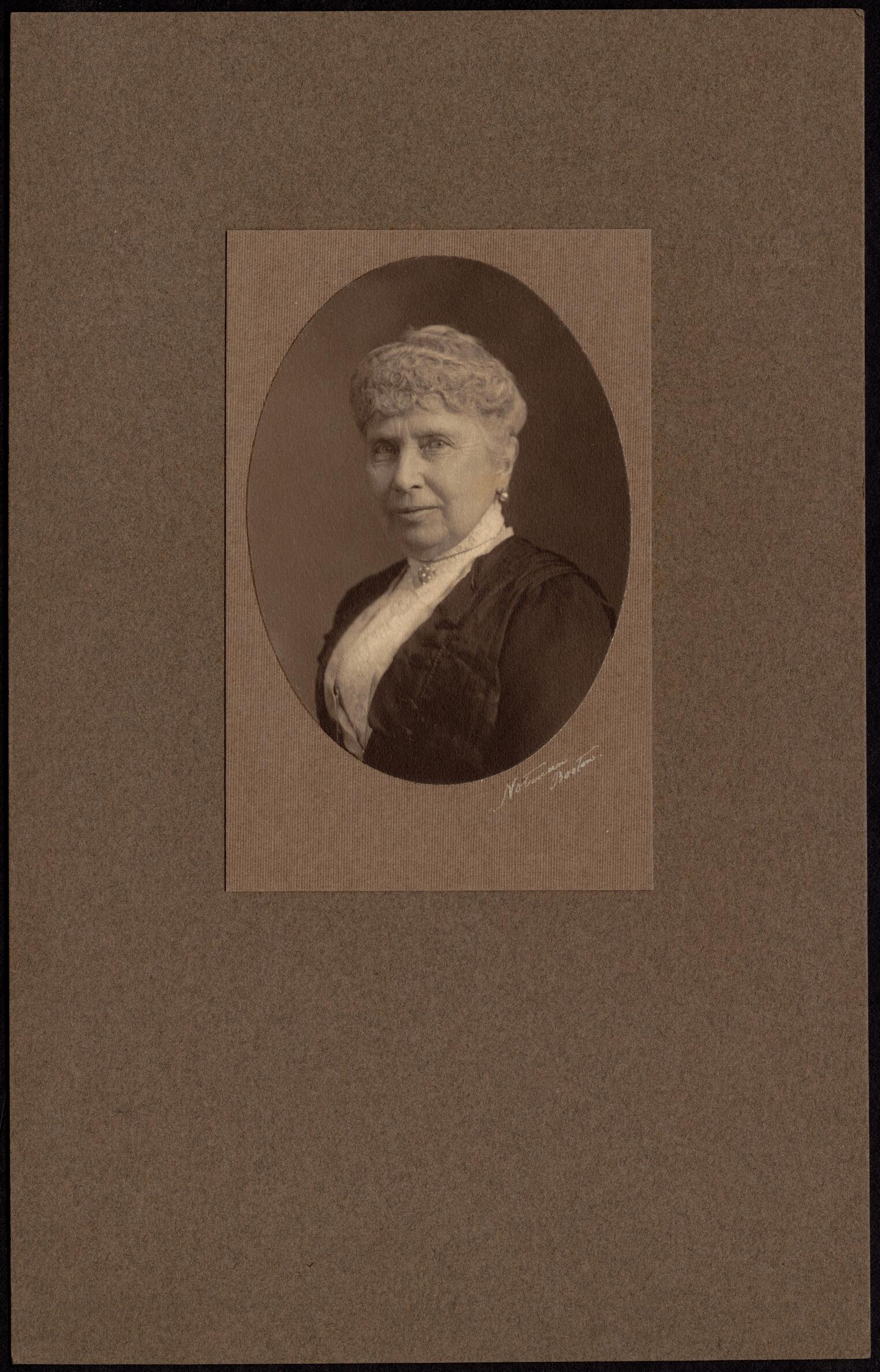 Brown-toned photographic portrait of a woman from the bust up, at an angle. She has light colored skin and gray hair in a curled style, and wears a dark dress with a high-collared white shirt.