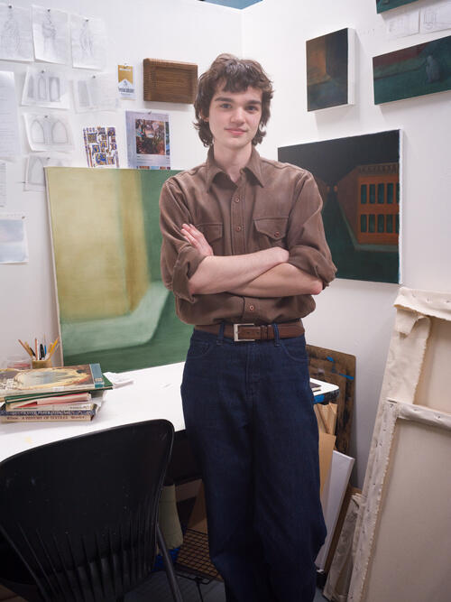A young white man standing with his arm crossed in front of him wearing a simple brown shirt and black pants