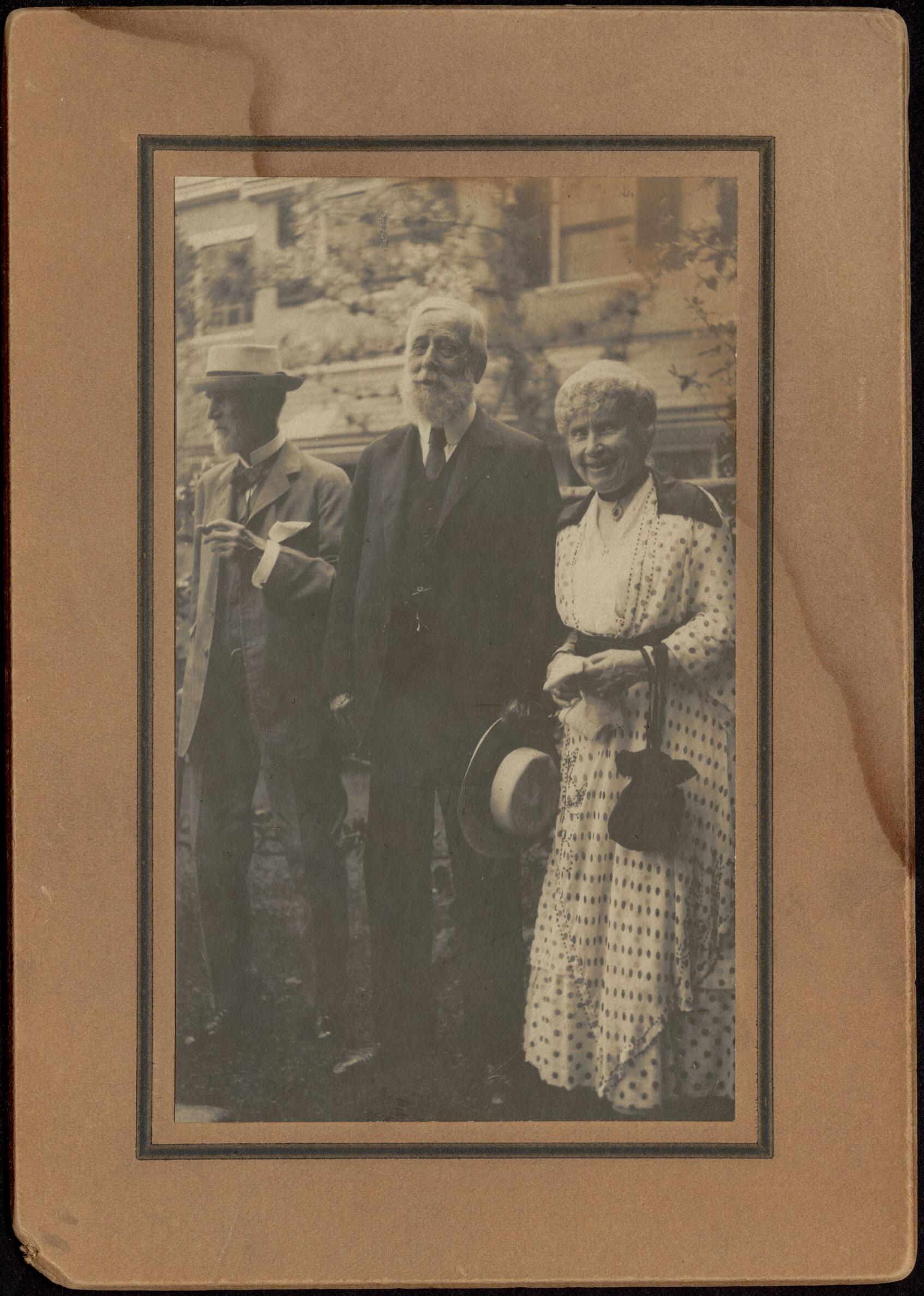 Brown-toned photograph of tree people standing in front of a building. The two men on the left wear dark suits and the woman on the right wears a light dress with a polka-dot pattern.