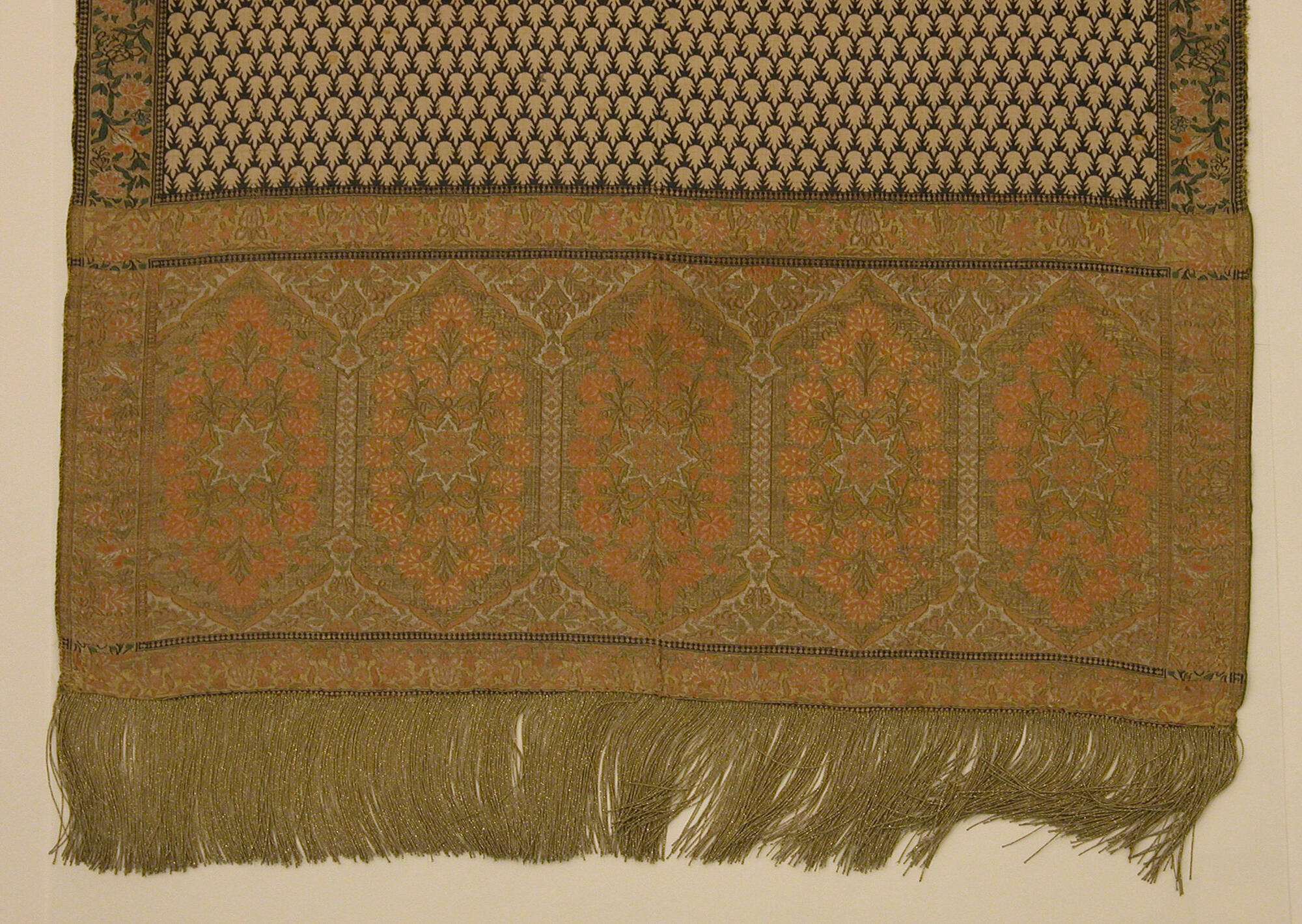 The end of a woven sash with intricate floral and geometric patterns in gold, coral, and dark blue with metallic fringe.