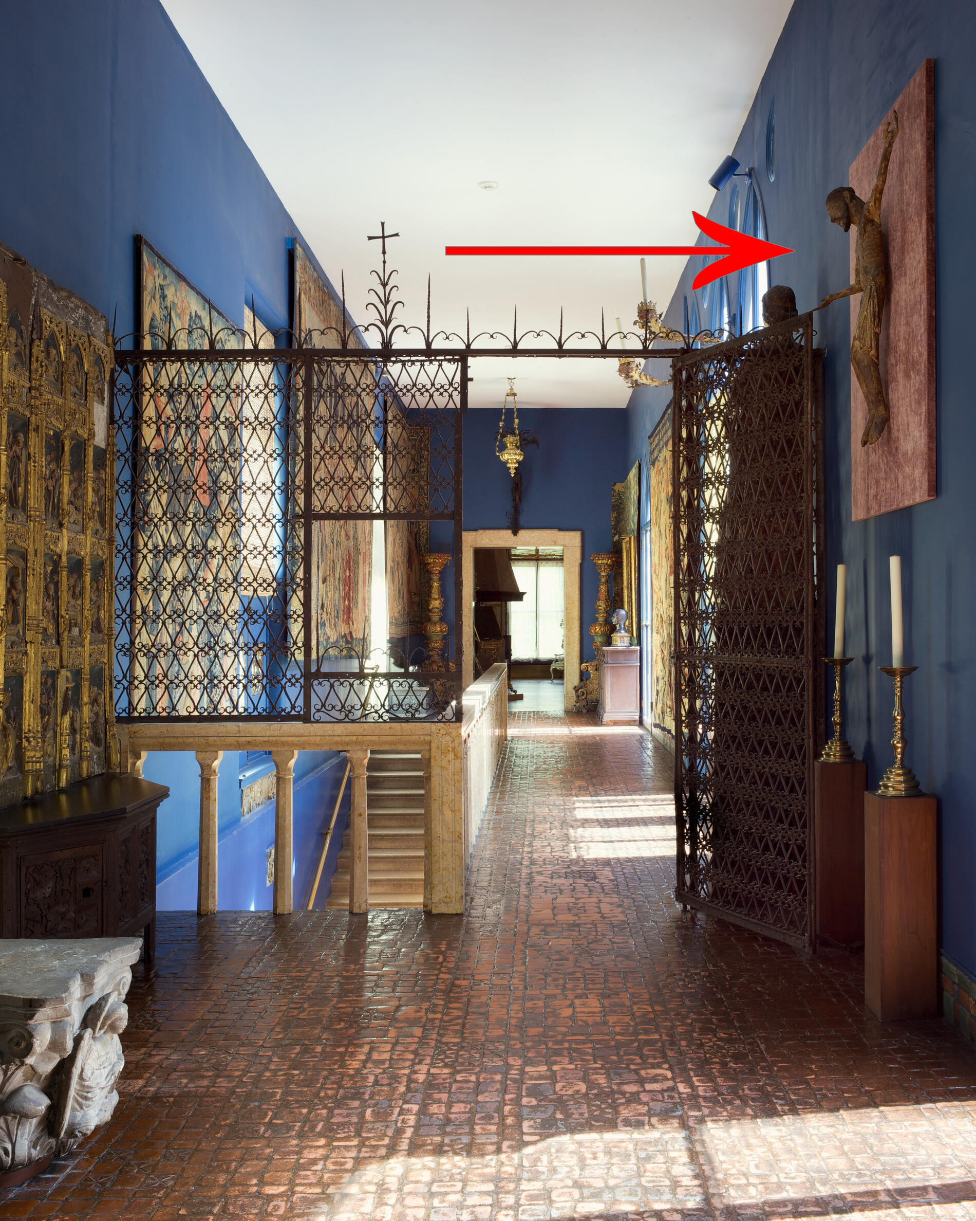 A stairhall with blue walls, an iron gate, and tapestries. A red arrow points to a wood sculpture of Christ hanging high on the wall.