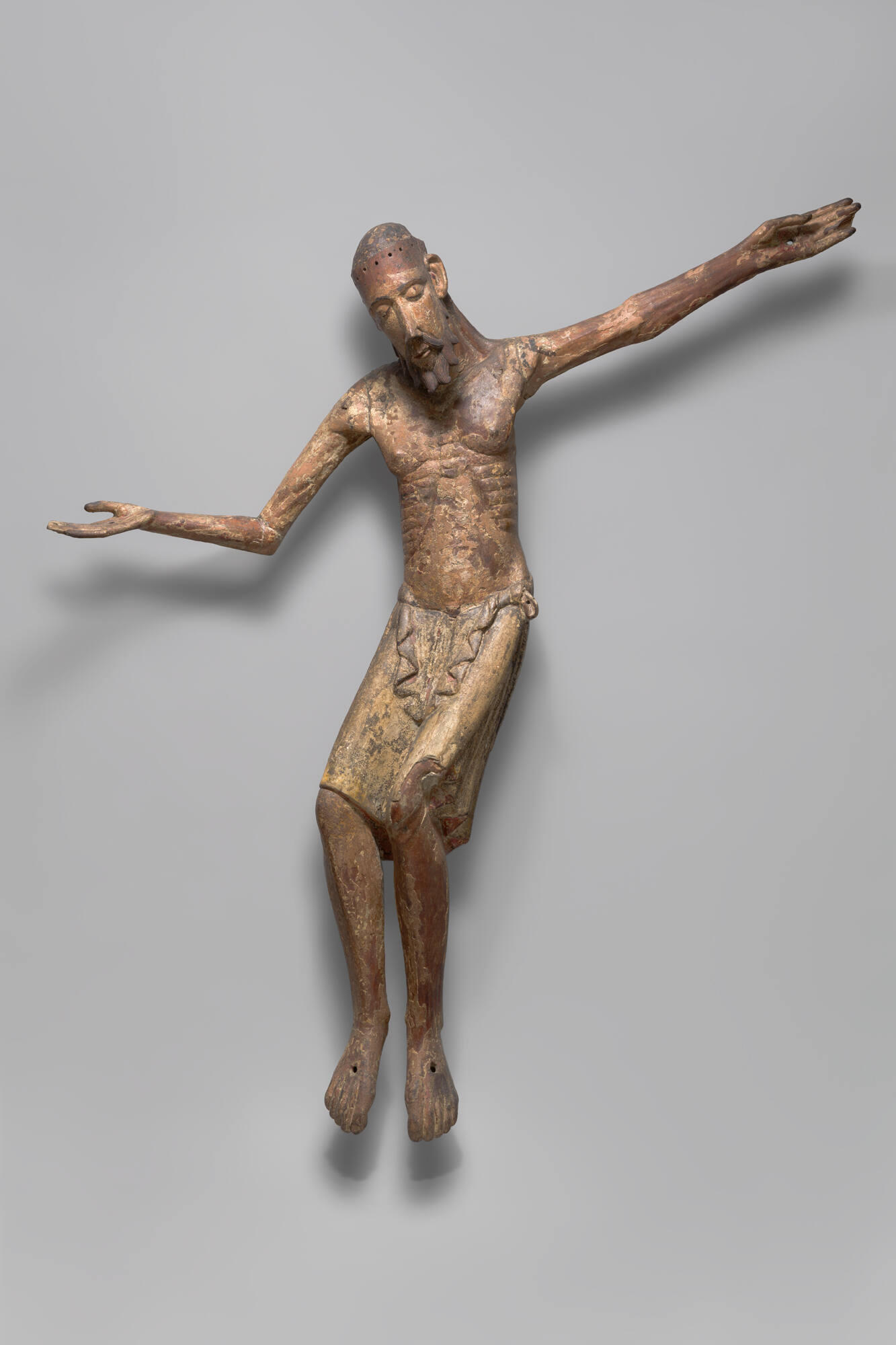 A painted wooden sculpture of Christ with eyes closed, dressed in a loincloth with ribs visible. His face has elongated features and a curled beard. One arm is raised and the other is pointed down. There are nail holes on his hands, with traces of painted stigmata.