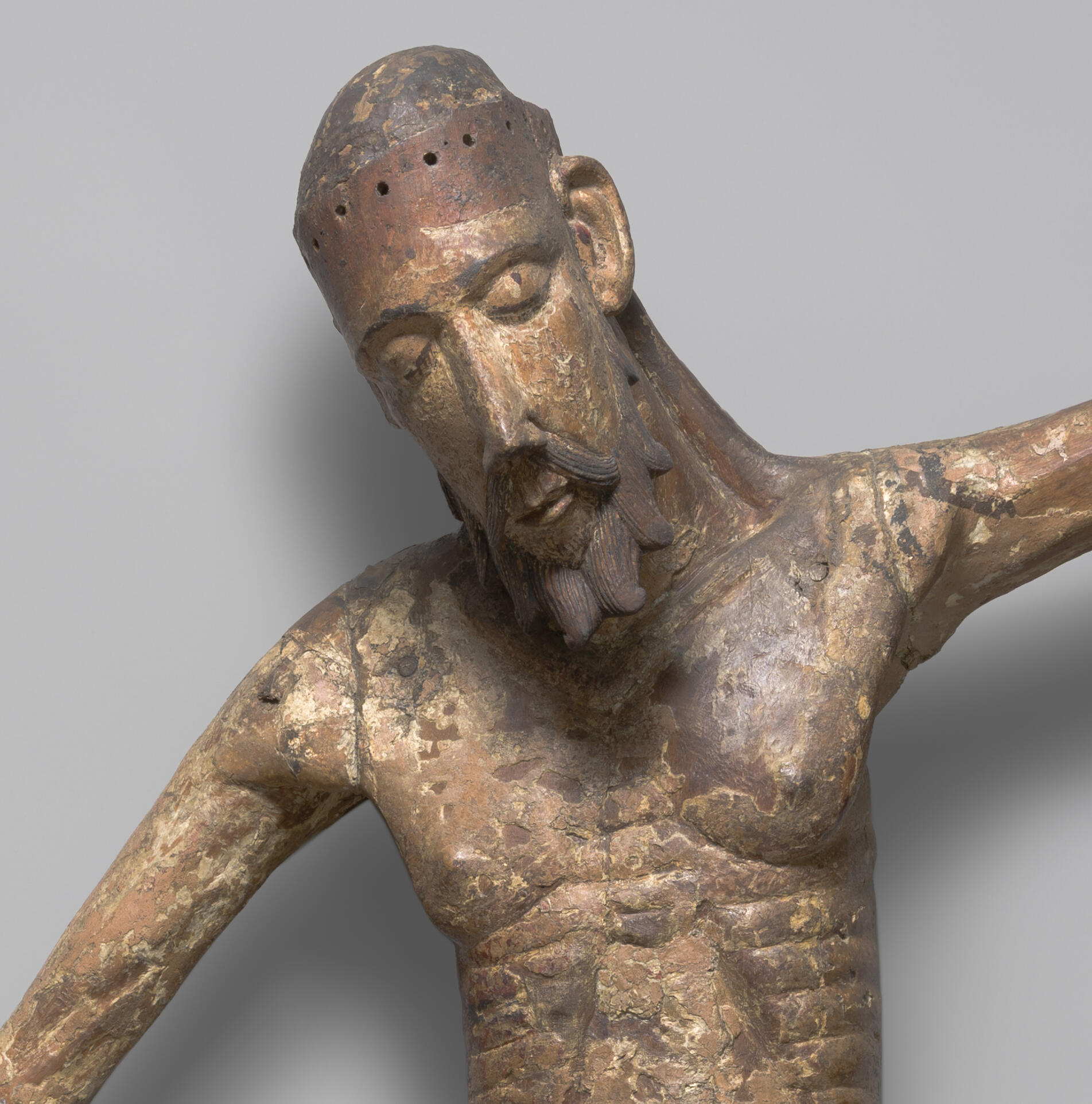 A wooden sculpture with traces of paint of Christ’s head and upper body. He has an elongated face and his eyes are closed. There are holes around his head where a crown of thorns would have been. His body is emaciated.