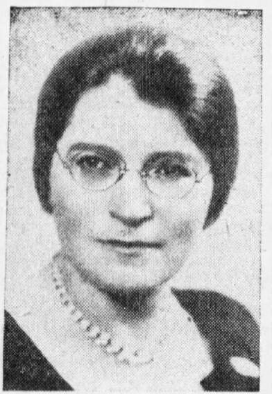 A black and white photo of the head and shoulders of a woman with dark hair wearing round wire-rimmed glasses, a necklace, and dark colored blouse or dress.