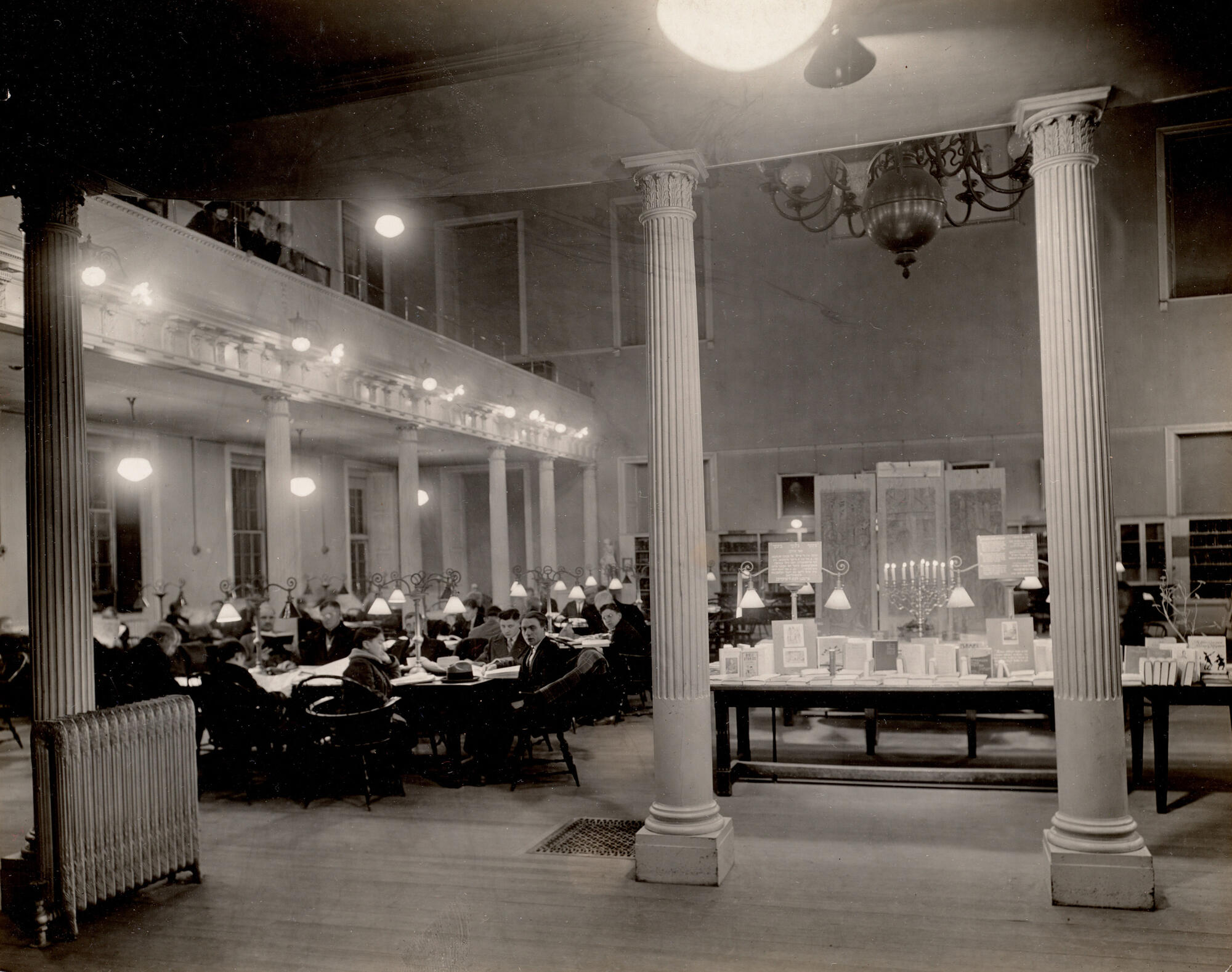A black and white photograph of a large room with people seated at tables with lamps looking at books.