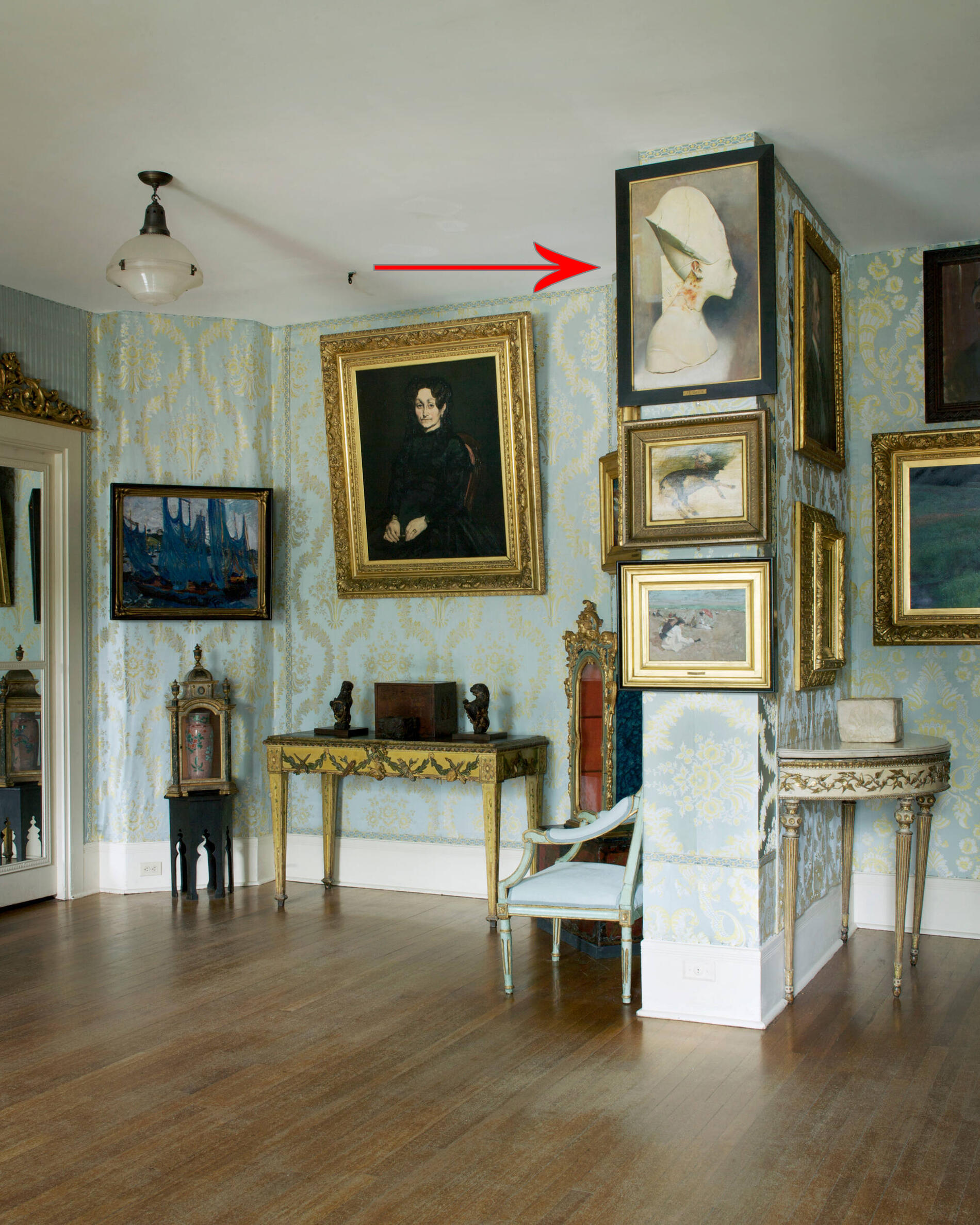 Wall with blue patterned wallpaper and many hung paintings, with tables, a chair, and cases sitting underneath them.