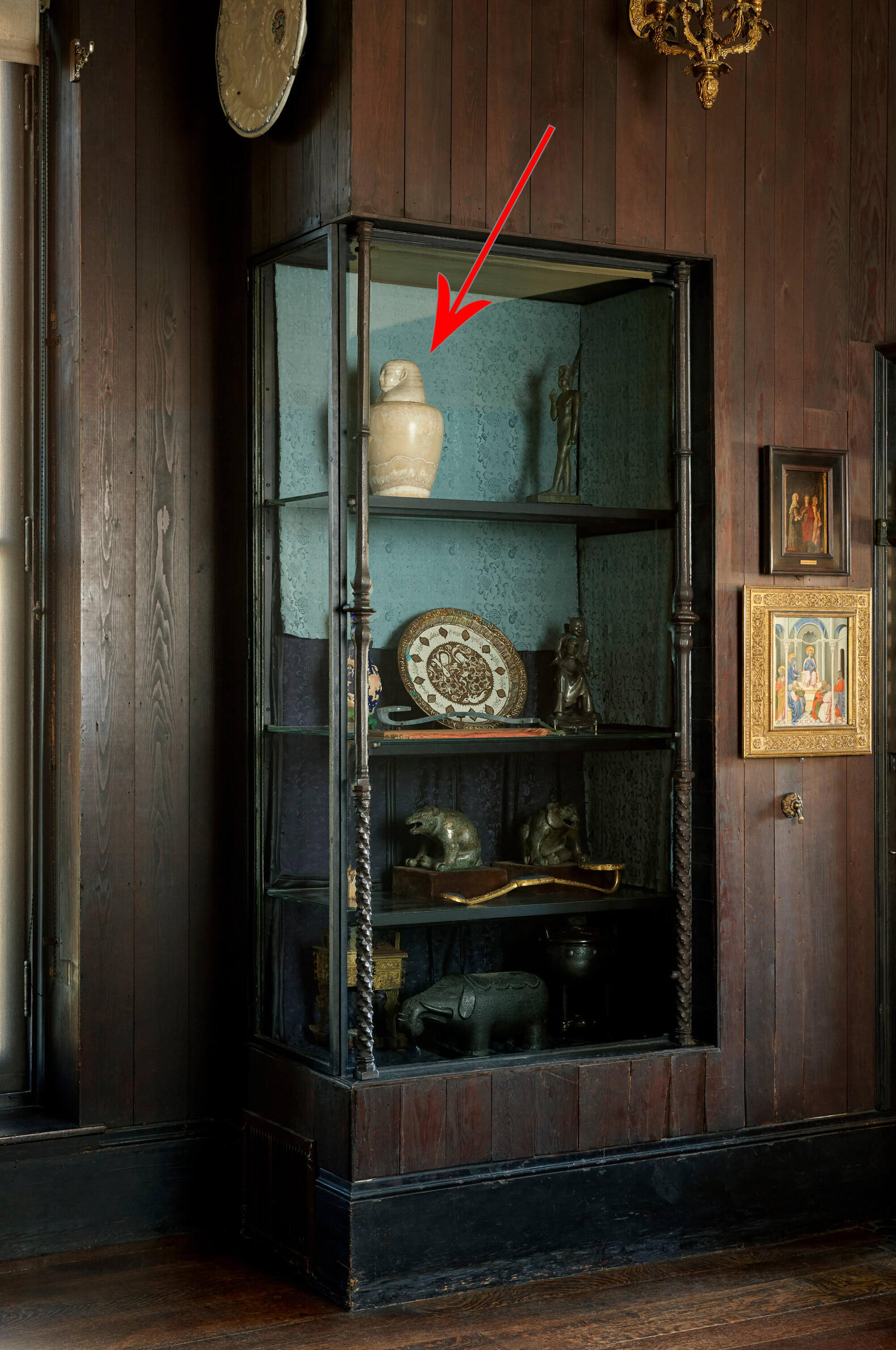 Glass case set against a wooden wall and filled with objects including sculptures, vessels, and a plate