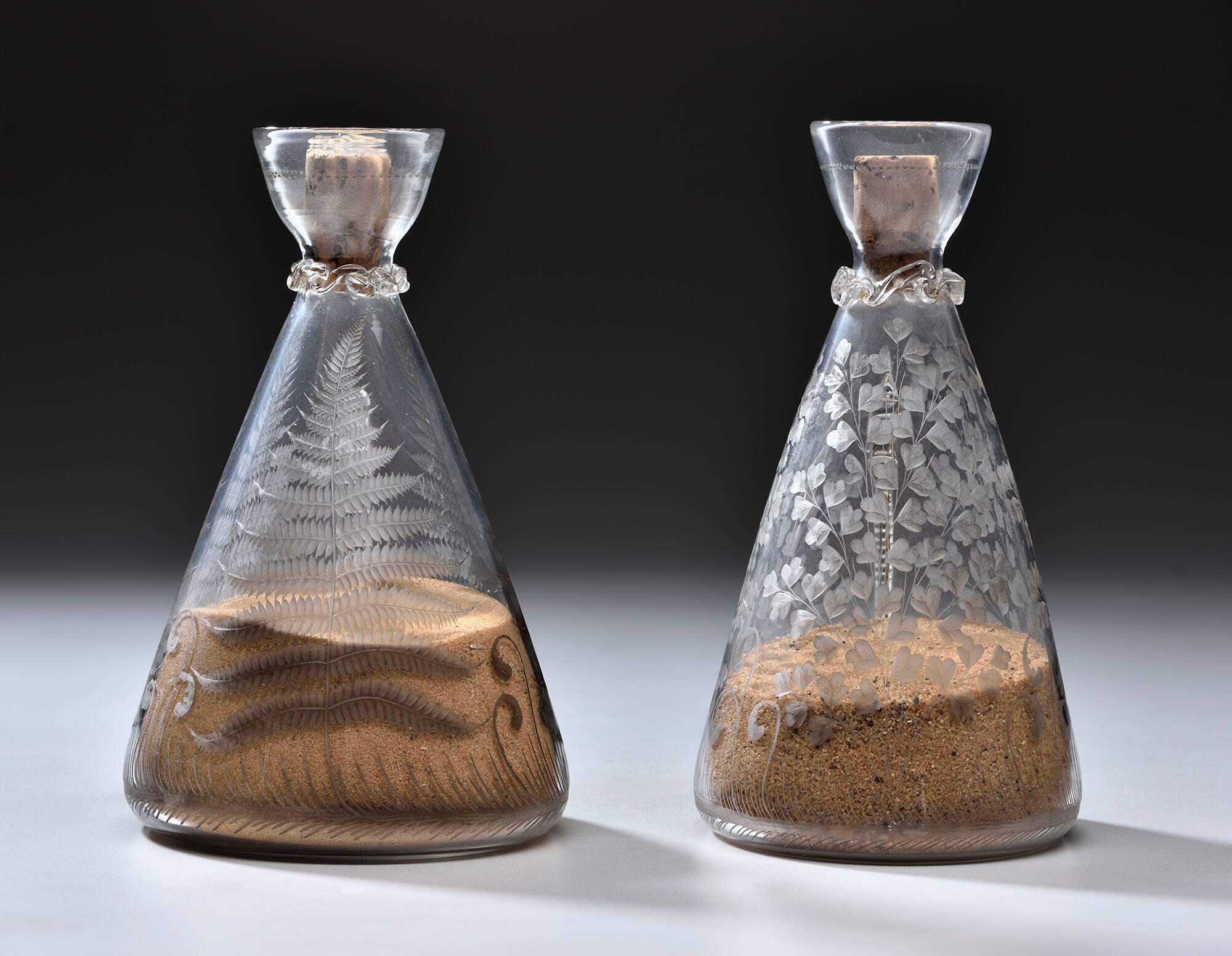 Two conical glass bottles with etched fern designs, filled partially with sand and corked.
