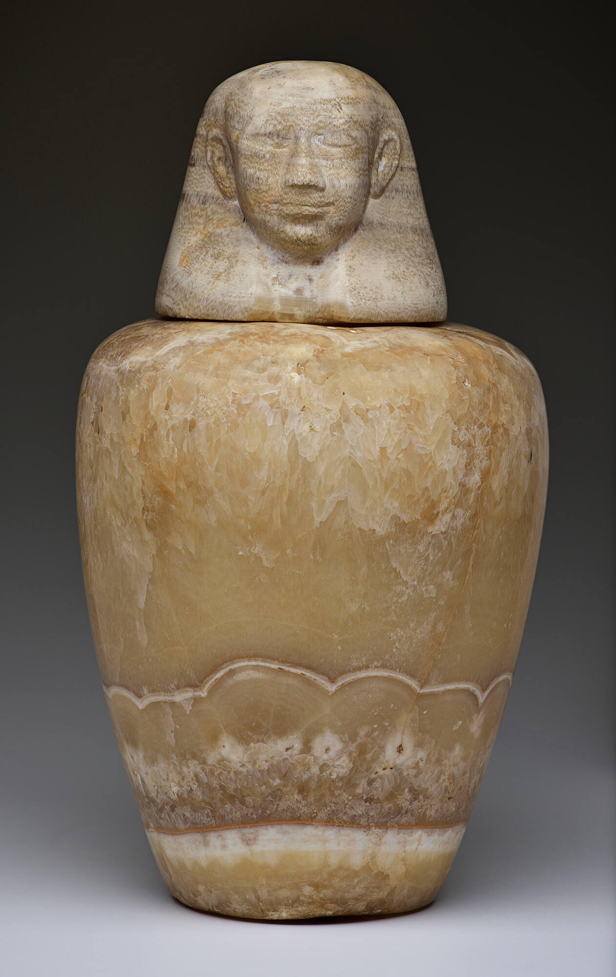 Tan jar with curved sides and a lid in the shape of a head. 