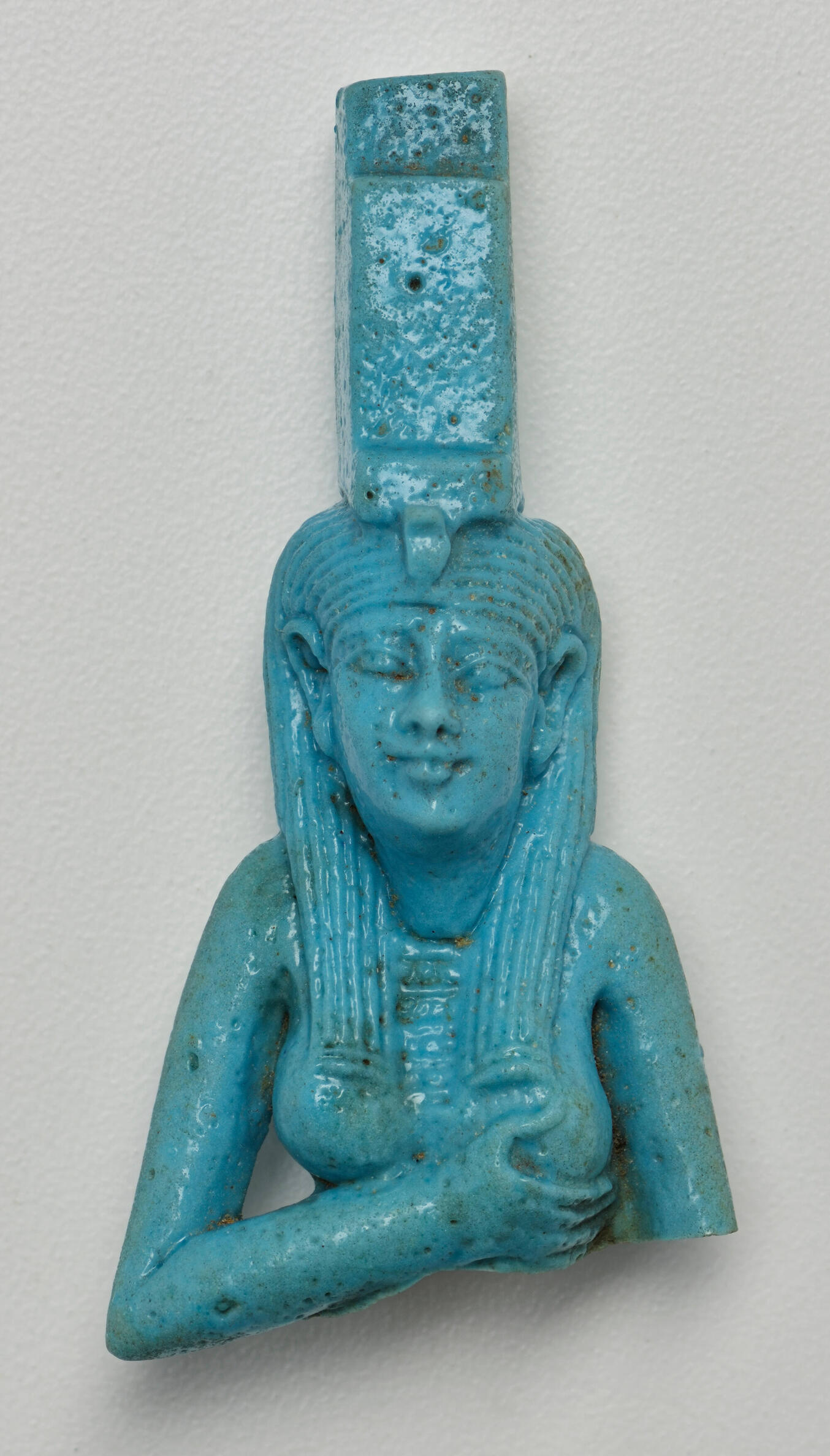 Fragment of a ceramic statue, glazed blue, depicting the chest, arms, and face of a female figure wearing a long, rectangular crown.