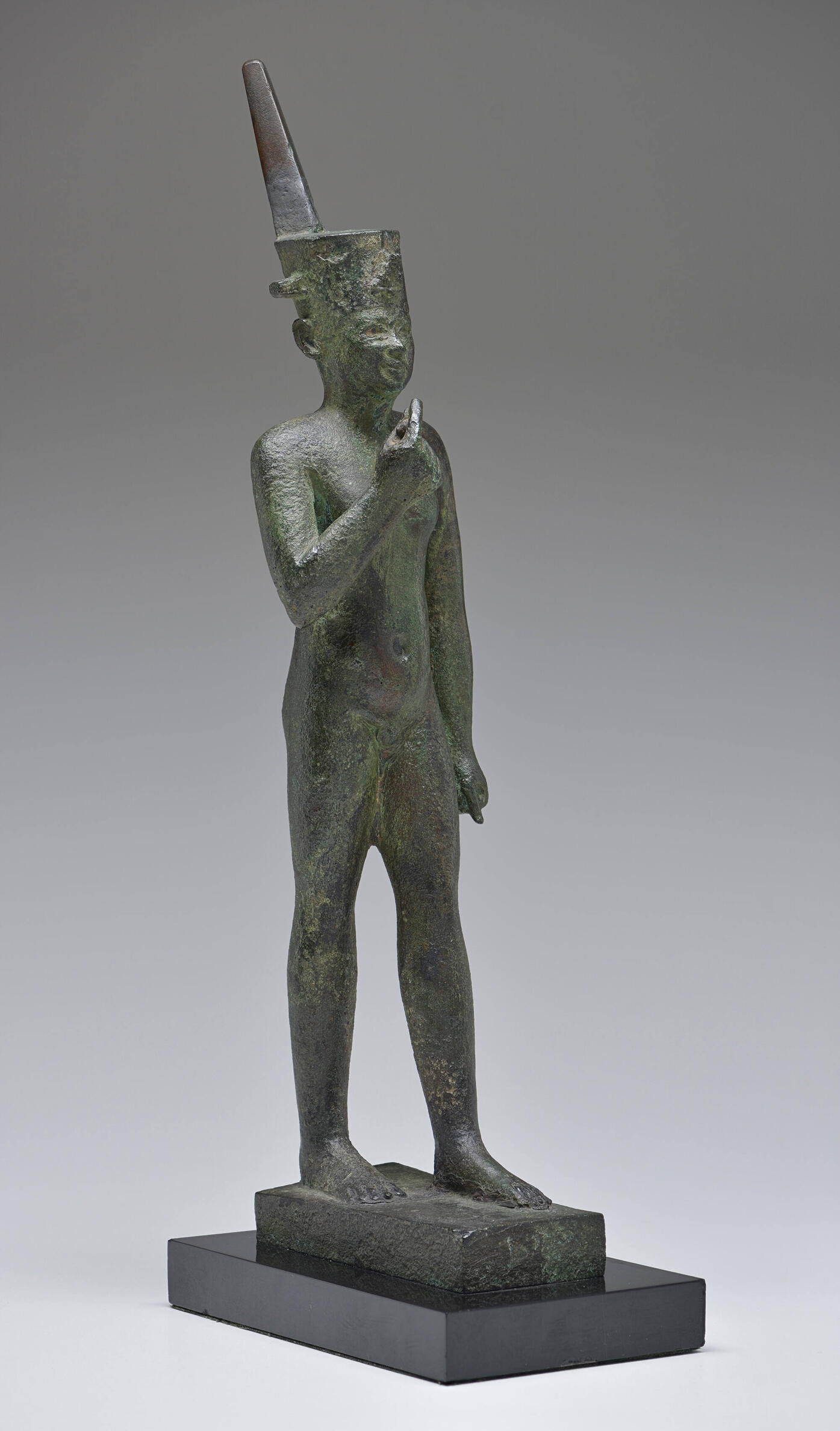 Bronze sculpture of a standing boy with one arm bent up, his finger pointing to his face.