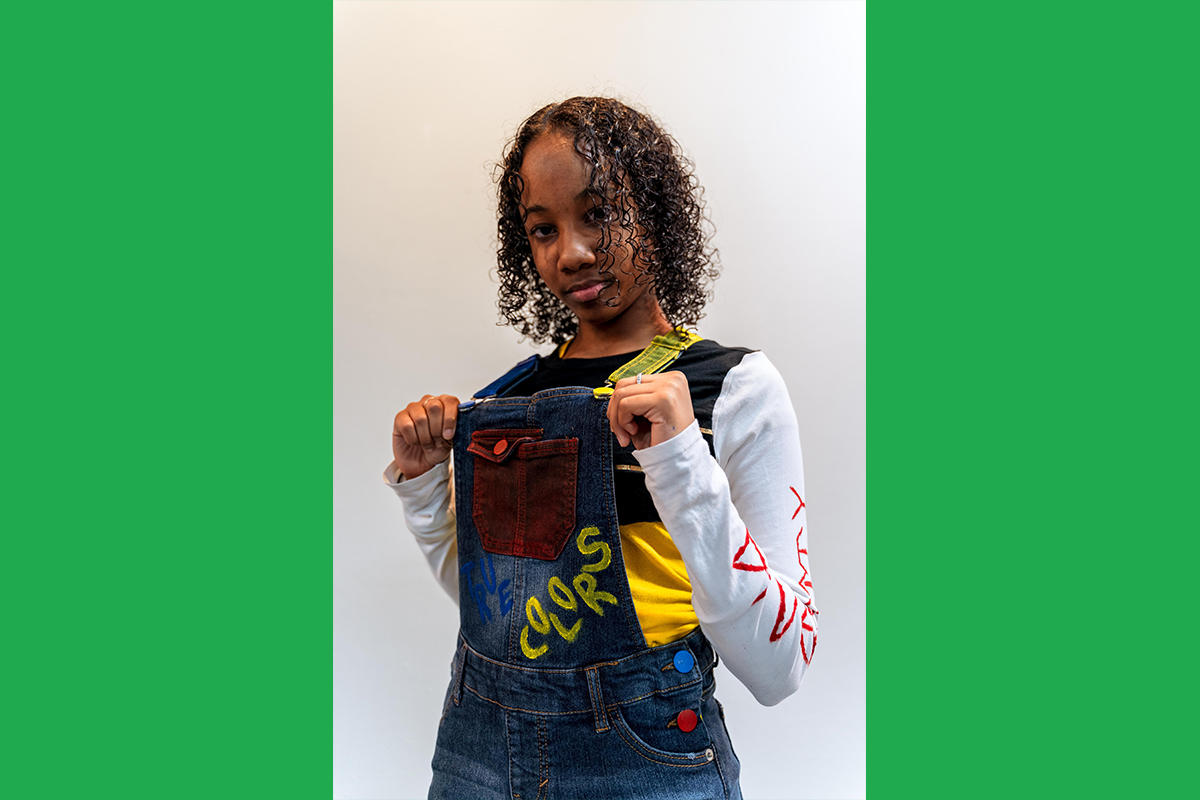 An Urbano Project student shows off their finished Map This garment: a pair of denim overalls with painted-over buttons and the word "colors" painted on the bib.